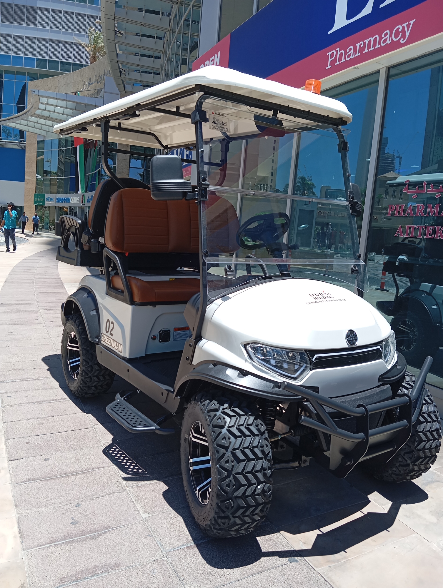 This is a full-shot, eye-level, outdoor photograph of a white, four-seater golf cart parked on a tiled walkway. The golf cart is the primary subject and is facing the viewer at a slight angle to the right. It has a white canopy, brown leather seats, and large, off-road tires. The number "02" and the word "GREENMAN" are visible on the side of the cart. The front of the cart has a black bull bar and "DUBAI HOLDING COMMUNITY MANAGEMENT" is printed on the hood. In the background, there is a modern building with large glass windows, and a sign that reads "Pharmacy" in English and Arabic. The overall environment suggests a commercial or residential area within Dubai, United Arab Emirates, possibly a community or resort given the presence of the golf cart and the branding. The walkway is made of light-colored square tiles. There are two individuals walking in the background on the left side of the image, dressed casually. The lighting indicates it is daytime with strong sunlight casting shadows.