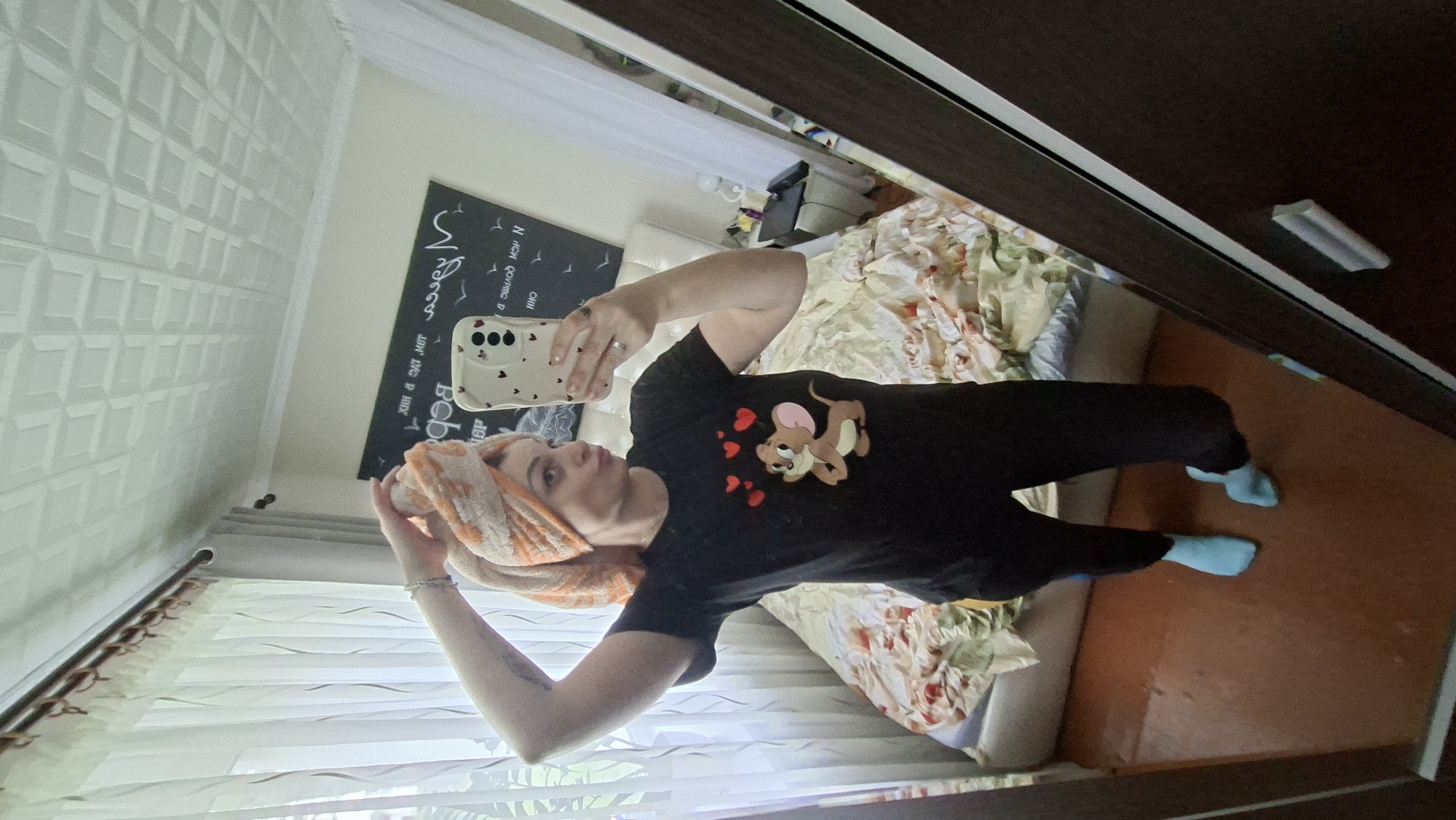 A woman is taking a mirror selfie in a bedroom. She is wearing a black t-shirt with a cartoon mouse and hearts printed on it, black leggings, and light blue socks. Her hair is wrapped in an orange and white towel. The room has a bed with a floral comforter, curtains, and a framed picture on the wall. The floor is a reddish-brown color. There is a mirror in the foreground reflecting the scene.