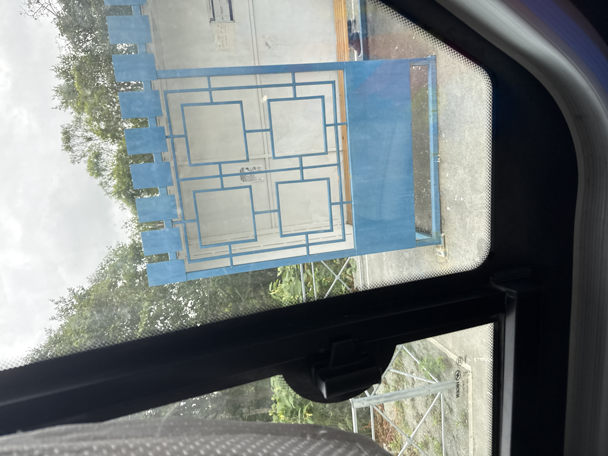 A bus stop shelter with a blue metal gate and a wooden bench is visible, with trees in the background and a cloudy sky. The shelter is located at a bus stop in Petropavlovskoe, Russia. The gate has a geometric design with square openings. A person is partially visible sitting on the bench, wearing dark pants and shoes. The view is through a window, with the frame of the vehicle visible in the foreground.