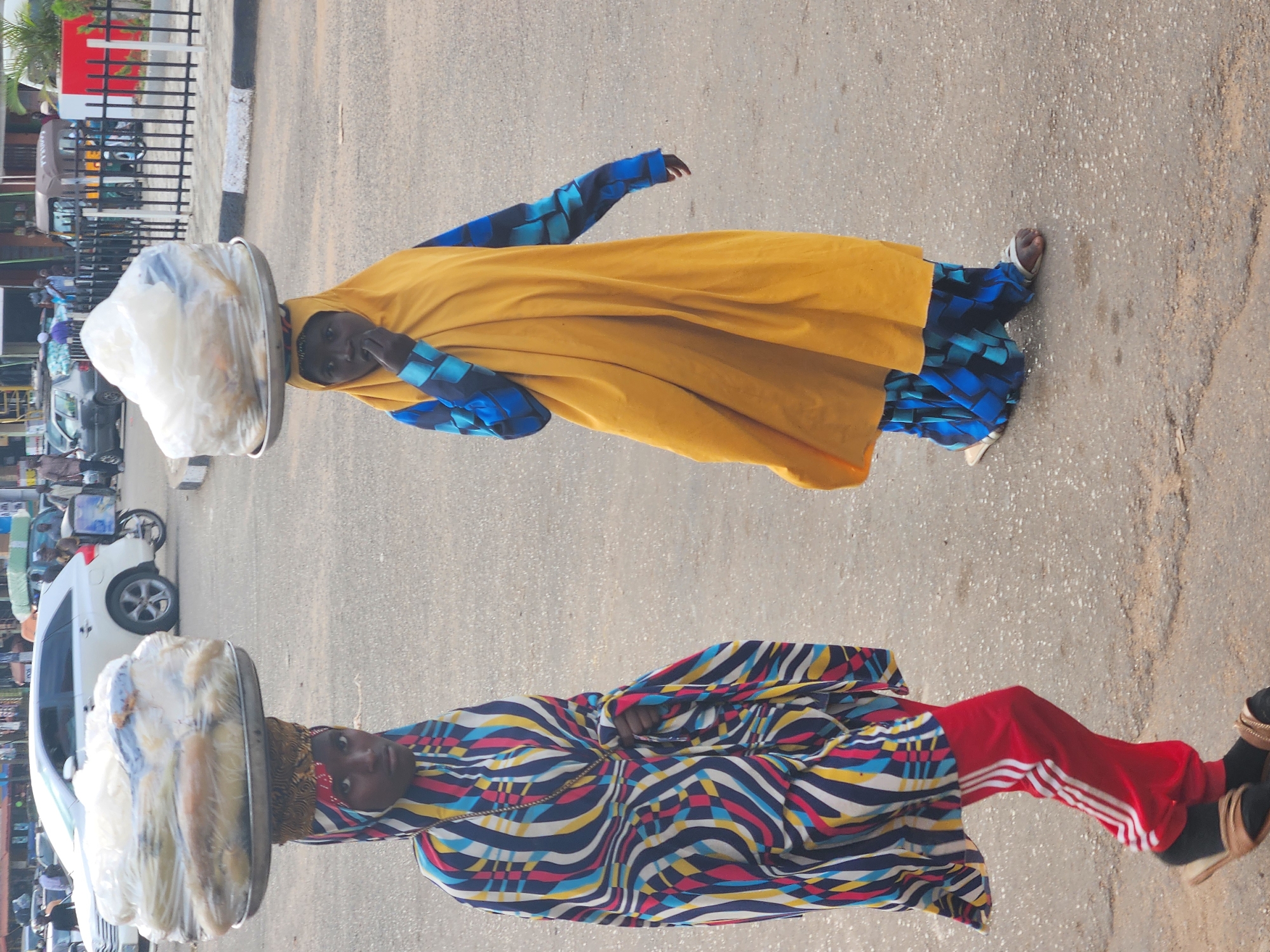 Two women are shown in an outdoor setting. One woman wears a yellow outer garment over a blue patterned inner garment, with a white object balanced on her head. The other woman wears a multicolored patterned top and red pants with white stripes. Both women appear to be standing on a paved surface. In the background are vehicles, including a white car, and various buildings.