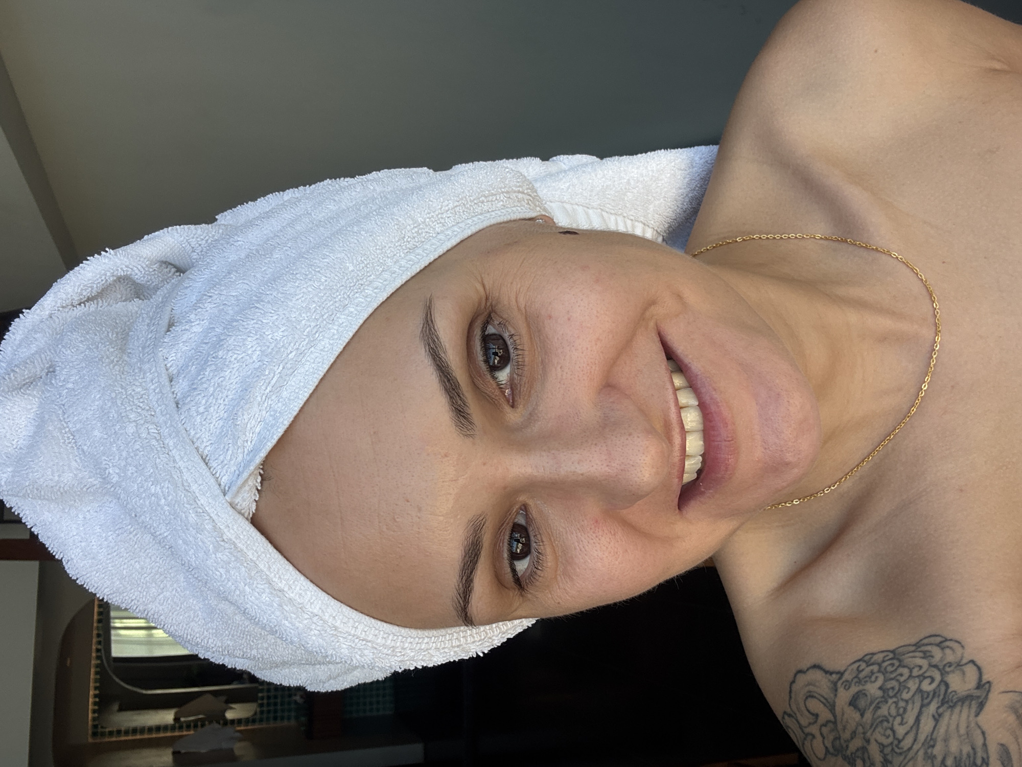 The image depicts a close-up, frontal view of a woman with a white towel wrapped around her head. She is bare-shouldered, wearing a thin gold chain. Her eyes are open, and she is smiling. A tattoo is visible on her left shoulder. The background is partially obscured by a dark area on the left, suggesting an indoor setting, possibly a bathroom or bedroom, with a hint of a mirror and tiled wall in the distance. The right side of the background appears to be a plain, neutral-colored wall. The lighting suggests a natural light source from the right. The image was likely taken indoors.