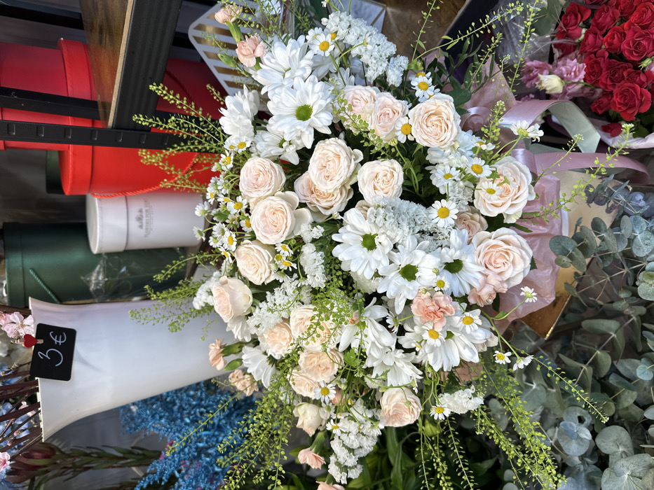 A detailed, close-up photograph depicts a large, full floral arrangement displayed within a commercial setting, identified as a florist shop in Liepaja, Latvia. The central subject is a voluminous bouquet primarily composed of light pink garden roses, white daisies with prominent yellow centers, smaller clusters of white filler flowers, and various green foliage, including delicate fern-like fronds. The bouquet is contained within a light-colored box, lined with pink tissue paper, with a pink ribbon tied around its lower portion.
In the background, to the upper left, a tall, white, flared vase is visible, to which a small black sign bearing "3€" in white script is clipped. Adjacent to this vase are bundles of blue-dyed, spiky botanical elements. Further back, an assortment of cylindrical storage or display boxes is present: several tall dark green boxes, a white box labeled "FLOWERS FOR YOU," and multiple red boxes, some stacked. A dark shelf with a wooden texture is partially visible on the upper right side of the frame. In the lower left foreground, broad, silvery-green eucalyptus leaves are visible. In the lower right foreground, partially obscured by the main bouquet, is a collection of dark red roses. No individuals are visible in the frame, and no specific actions or interactions are depicted beyond the static display of floral products.