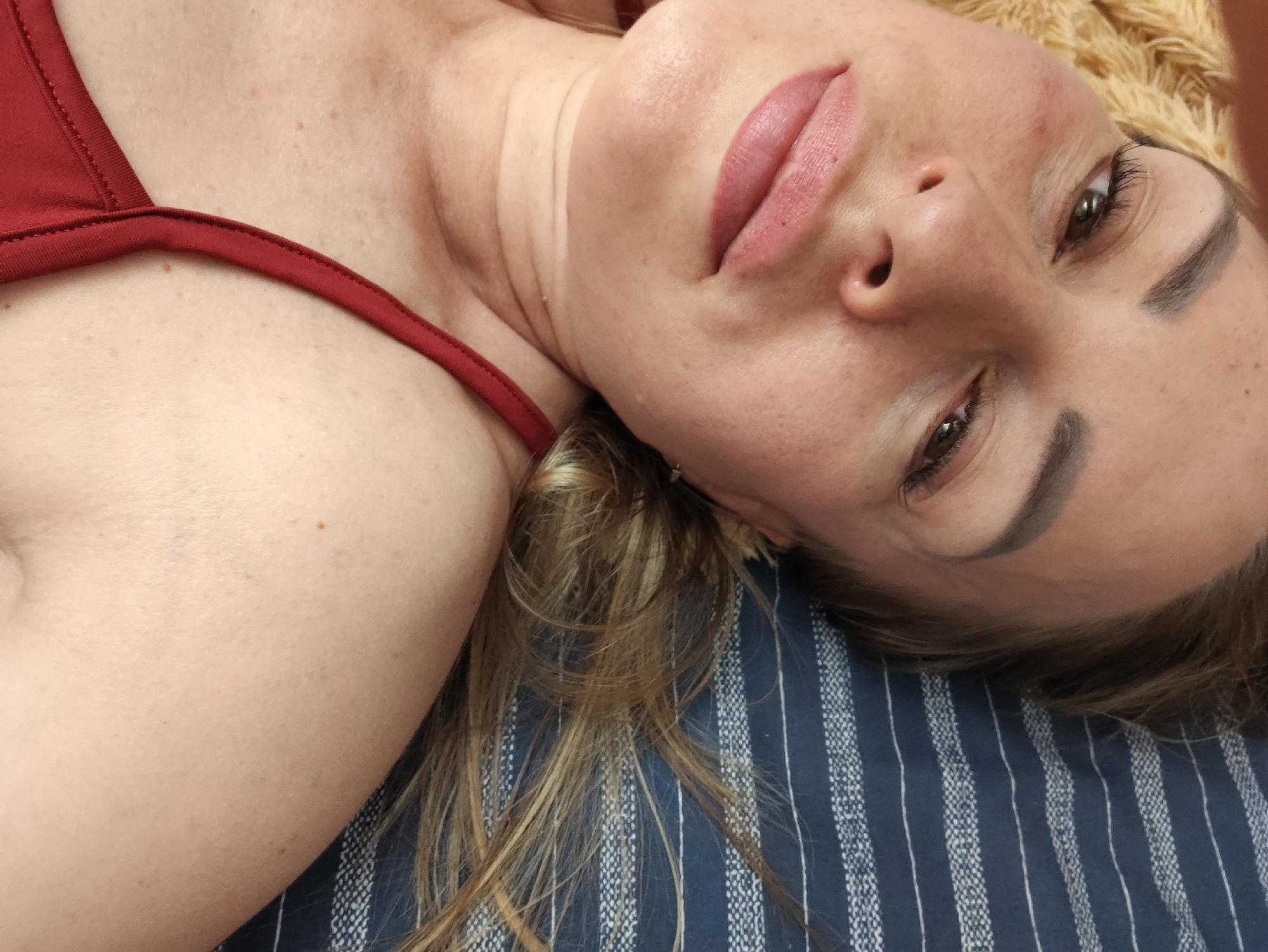 A close-up, top-down, over-the-shoulder shot shows a woman lying on a striped blue and white surface with a tan towel behind her head. She is wearing a red garment. Her head is tilted to her right, with her face turned toward the camera. Her lips are slightly parted and are adorned with pink lipstick. Her left eye is visible and looking away from the camera, while her right eye is partially obscured. The lighting casts soft shadows on her face, highlighting her cheekbones and the contours of her neck. Her blonde hair is spread across the surface. The image does not provide any information about the location in Almaty, Kazakhstan.