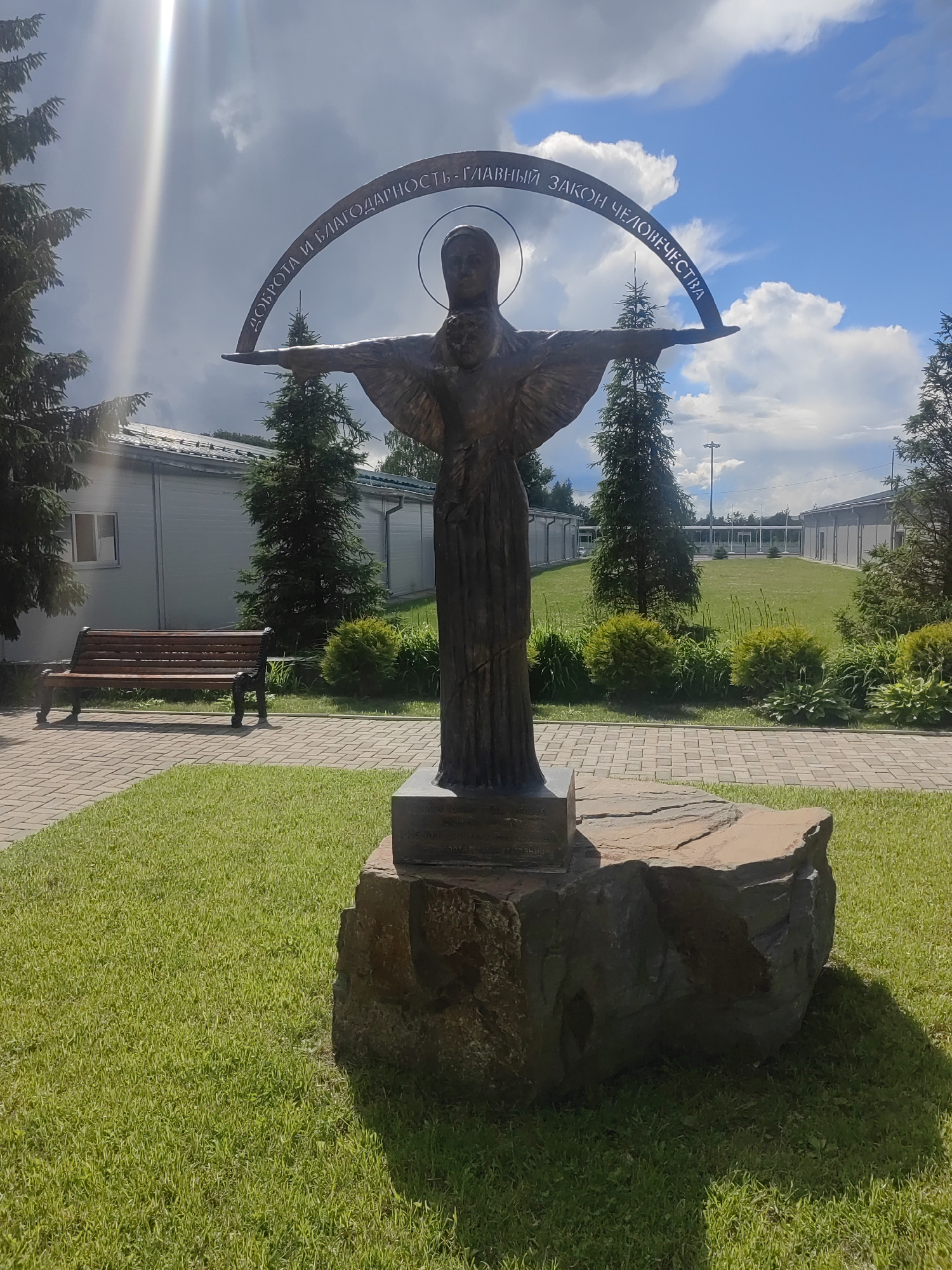 A bronze sculpture depicts a winged figure with outstretched arms, holding a crescent-shaped banner above its head. The banner is inscribed with Cyrillic text. A halo encircles the figure's head. The sculpture stands on a large, rough-hewn stone pedestal, which is placed on a paved pathway.
The sculpture is situated in an outdoor setting. To the left of the sculpture, a wooden bench with dark metal legs sits on a brick pathway surrounded by a neatly mowed green lawn. Behind the bench and to the left of the sculpture, a modern building with light-colored walls and a grey roof is visible, with windows and fir trees in front of it. To the right of the sculpture, more fir trees and manicured bushes are visible, with similar modern buildings in the background. The scene appears to be a park or grounds of an institution.
The time of day is daytime, indicated by the bright sunlight casting shadows. The weather appears to be partly cloudy, with dramatic sunbeams breaking through the clouds, suggesting fair weather with some cloud cover. The inscription on the banner reads: "ДОБРОТА И БЛАГОДАРНОСТЬ – ГЛАВНЫЙ ЗАКОН ЧЕЛОВЕЧЕСТВА" which translates to "Kindness and Gratitude are the Main Law of Humanity." The overall mood is serene and contemplative, emphasizing the message of the sculpture. The location context is Kubinka, Russia.