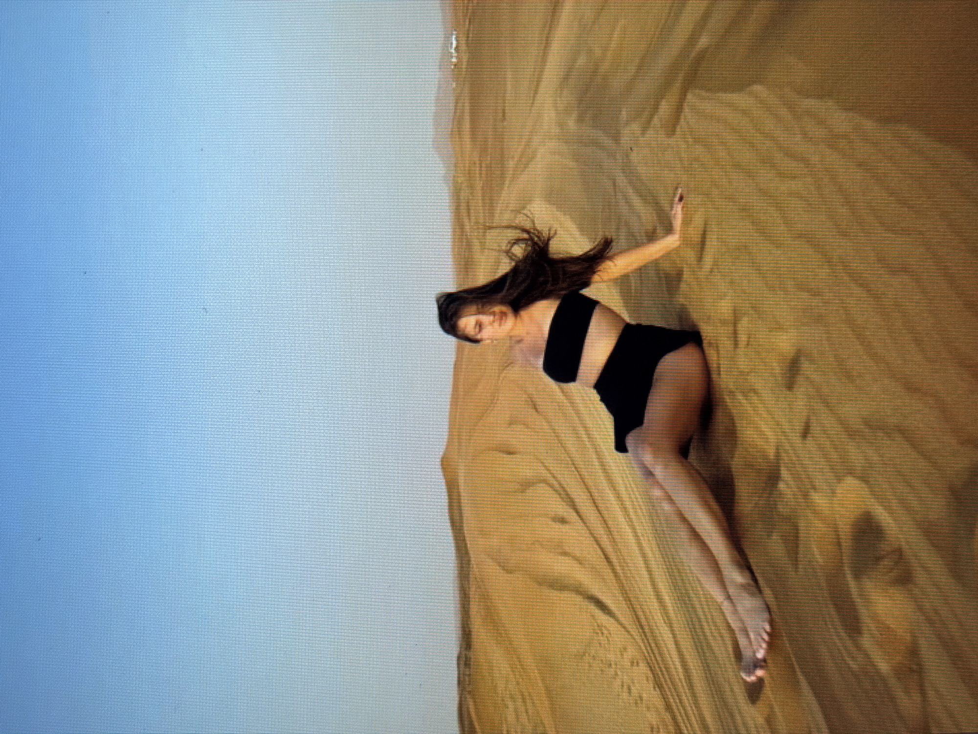 The image shows a young woman in a black bikini sitting on a sand dune. She is leaning back on her left hand, with her legs extended in front of her. Her hair is long and dark, and it is blowing in the wind. The background is a vast expanse of sand dunes, with a clear blue sky overhead. There are faint tracks visible in the sand, suggesting that a vehicle may have passed through the area. In the distance, there are some buildings visible on a hill. The location is identified as the city of Sirius, Russia, which is a resort city located on the Black Sea coast.