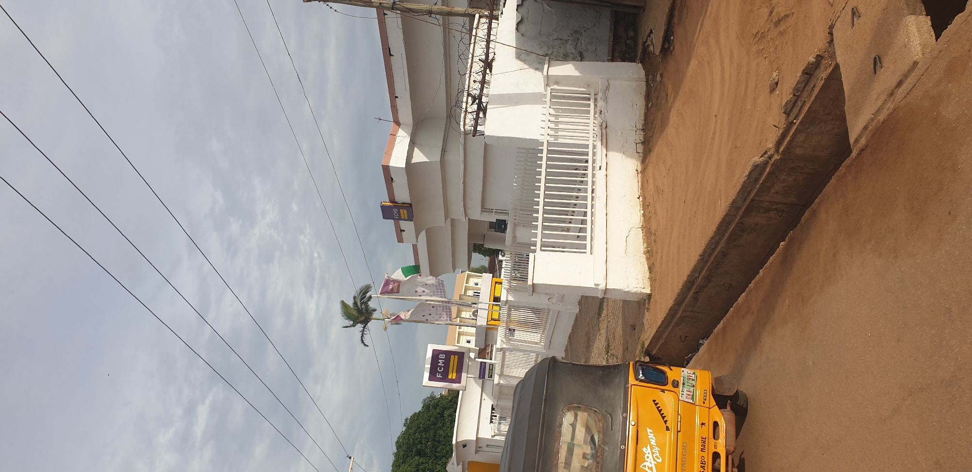 The image captures a street scene in Sabon Tasha, Nigeria, under an overcast sky. Dominating the foreground is the rear portion of a yellow, three-wheeled auto-rickshaw, identified by markings like "Ape City" and "VIAGGIO". Its license plate is visible, reading "OKA - 129VE". To the right of the auto-rickshaw, a concrete drainage channel runs along the side of a dirt road.
In the middle ground, a white building with a brown trim is prominent, identified by "FCMB" signage on a purple rectangle and also on the building's facade. Two flags, one white with a green stripe and another with a Nigerian flag, are displayed in front of the building. A white metal fence runs along the front of the property. Behind this building, another white structure with yellow accents is visible. To the far left, a lush green tree stands tall. The background is characterized by utility lines crisscrossing the sky. The overall impression is a typical urban or semi-urban setting in Nigeria.