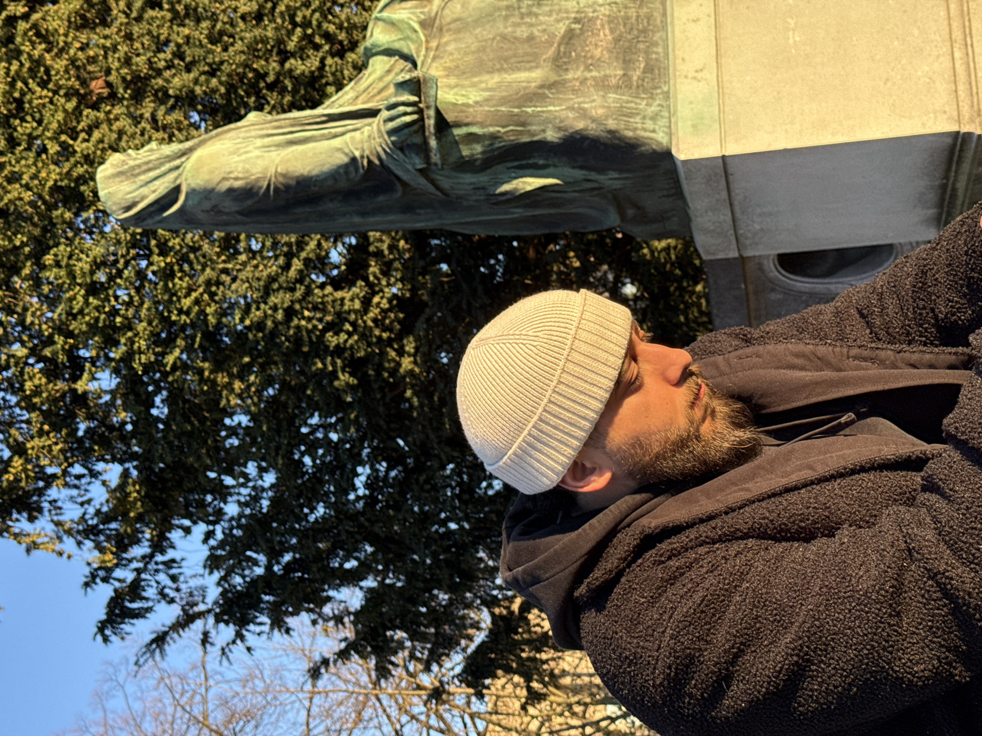 The image captures a male individual in the foreground, situated outdoors under clear blue skies in Place Kléber, Strasbourg, France. The subject, seen from the chest up and facing downwards, wears a light-colored ribbed beanie and a dark, textured jacket over a dark hooded sweatshirt. His face, partially obscured by his posture, is illuminated by golden, low-angle sunlight, highlighting his beard. To his right and slightly behind him stands a large bronze statue with a greenish patina, depicting a robed, hooded figure seated or kneeling and facing right. This statue represents "Alsace Mourning Her Son," part of the monument to General Kléber. The statue rests on a light-colored stone base. The background features dense green foliage of evergreen trees bathed in sunlight, with some bare deciduous tree branches visible to the left. The scene is lit by strong, warm sunlight, indicating either early morning or late afternoon.