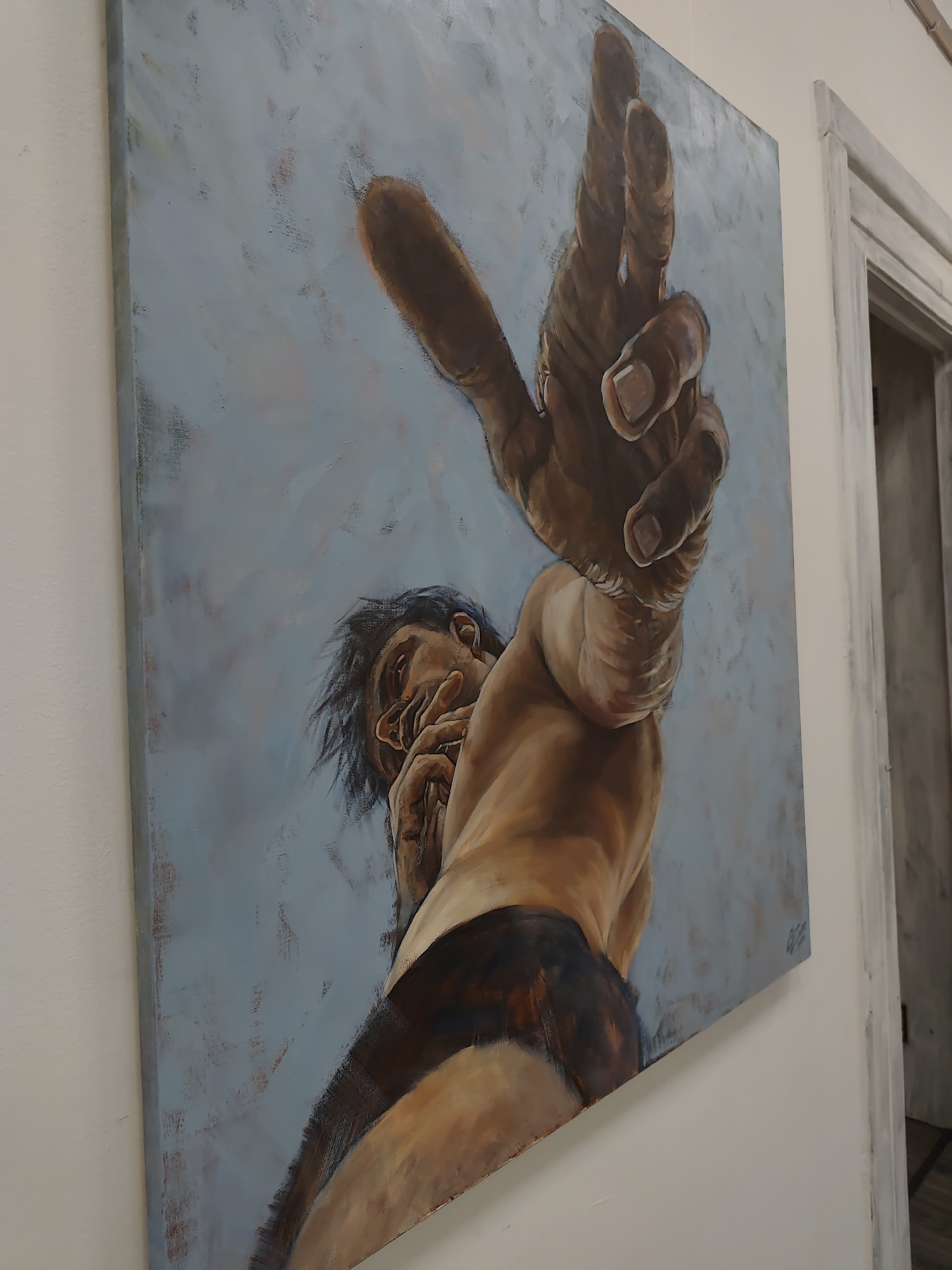 This is a photograph of a painting hanging on a wall. The painting is an oil on canvas, depicting a person from a low angle, looking up. The subject has dark hair and appears to be wearing dark briefs. Their torso is exposed, and their skin tones are rendered in warm browns and tans, contrasting with the cool, muted blue background. The most prominent feature is the subject's raised hand, extending towards the viewer, with fingers spread as if reaching out or pointing. The subject's face is partially obscured, with their hand near their mouth, suggesting an introspective or thoughtful pose. The brushstrokes are visible, giving the painting a textured, somewhat raw quality. The lighting in the photograph suggests an indoor setting with artificial light. To the right of the painting, a portion of a doorway with a distressed, light-colored frame is visible, indicating the painting is displayed in a room or hallway. The location context is Sochi, Russia. There is a faint signature in the lower right corner of the painting, but the text is illegible.