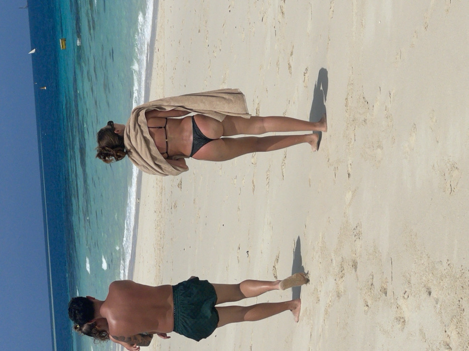 Two people are on a sandy beach. One is a woman facing the ocean, wearing a dark bikini and draped in a light brown towel. The other is a man, shirtless and in dark green shorts, facing the opposite direction. The turquoise ocean is in the background. The scene appears to be taken during a sunny day as evidenced by the shadows.