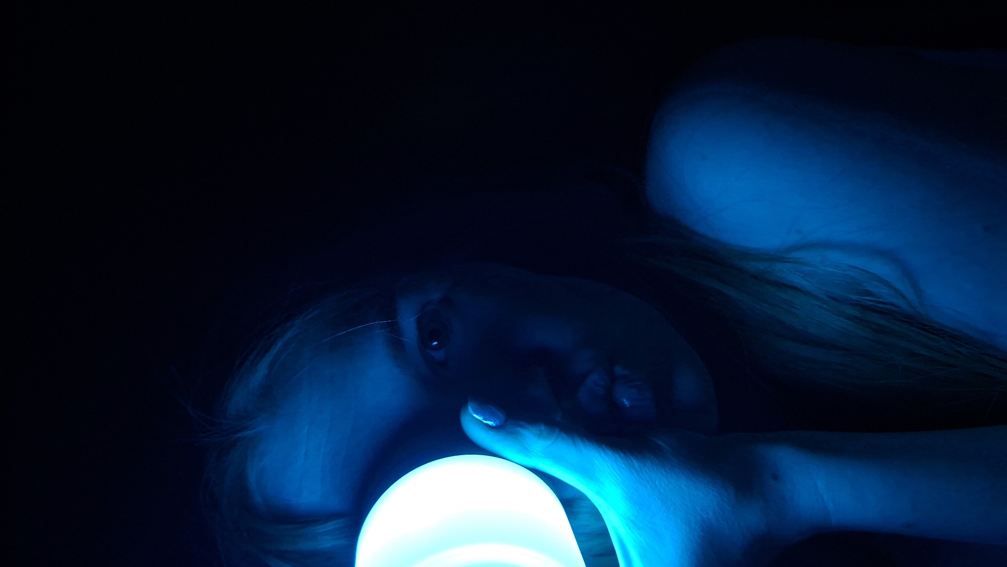 A person is holding a glowing blue orb in their hand. The person has blonde hair and fair skin, and the face is illuminated with a blue light. The background is dark, with only parts of the person's shoulder and torso visible. The image gives no indication of being in the city of Chaikovsky, Russia.