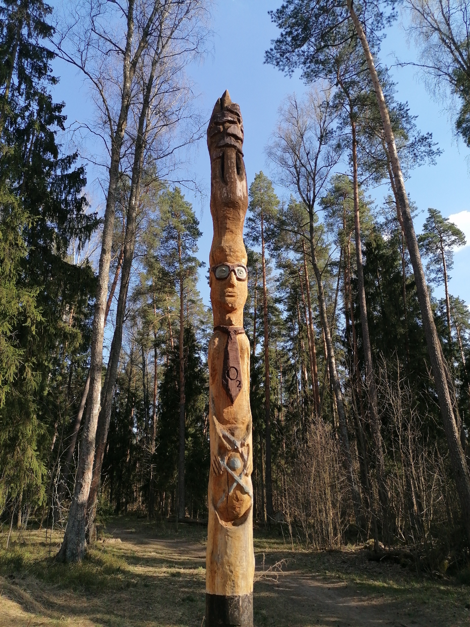 A tall, carved wooden totem pole stands in a grassy clearing within a forest. The totem features a face with eyeglasses, a necktie with "O2" written on it, and a symbol lower down on the pole. The pole is surrounded by tall trees, with a blue sky visible overhead. The ground is covered with grass and some fallen leaves, and a dirt path is visible nearby.