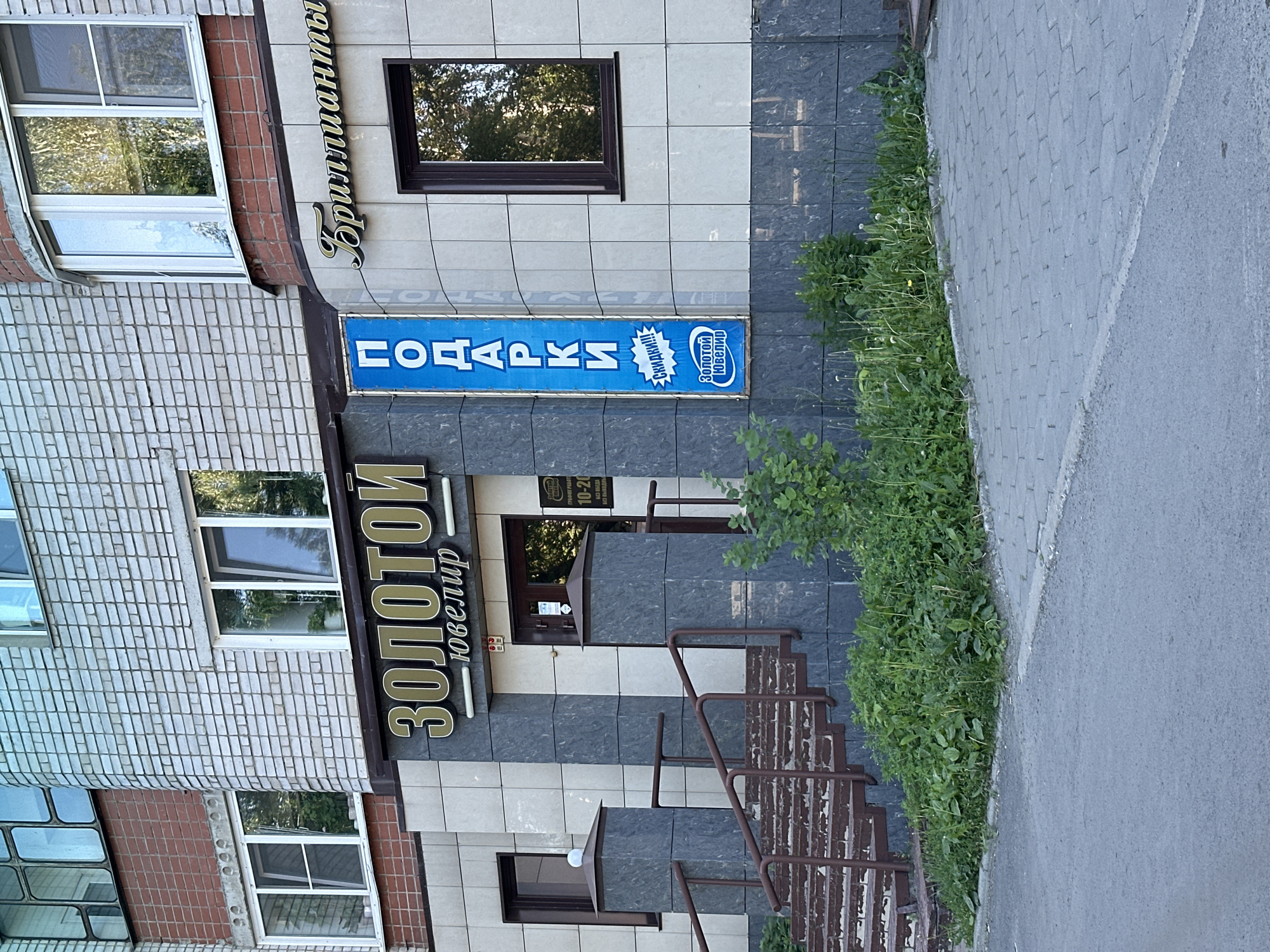This image shows the exterior of a jewelry store called "Золотой Ювелир" (Golden Jeweler) located in the city of Novoaltaysk, Russia. The store is situated on the ground floor of a multi-story building constructed with light-colored brick and beige tiled facade. A set of brick stairs with brown metal railings leads up to the store's entrance. The entrance features a double glass door with a sign indicating operating hours and a small logo.
Above the entrance, a prominent sign in gold lettering reads "ЗОЛОТОЙ Ювелир". To the right of this sign, a curved section of the building facade has a sign in stylized script that says "Бриллианты" (Diamonds). Adjacent to the "Золотой Ювелир" sign, and to the left of the entrance, is a vertical blue banner with white lettering that spells out "ПОДАРКИ" (Gifts). Below this, a smaller graphic in a blue oval indicates "Скидки!!!" (Discounts!!!) and another blue oval contains the text "Золотой Ювелир".
The building has several windows visible, some with reflective glass showing trees and sky. The area in front of the store is paved with gray interlocking concrete pavers, with a strip of asphalt in the foreground. Lush green vegetation and some weeds grow alongside the stairs and the base of the building, with some dandelion flowers visible. The lighting suggests it is daytime, likely a sunny day, casting some shadows. There are no people visible in the image.