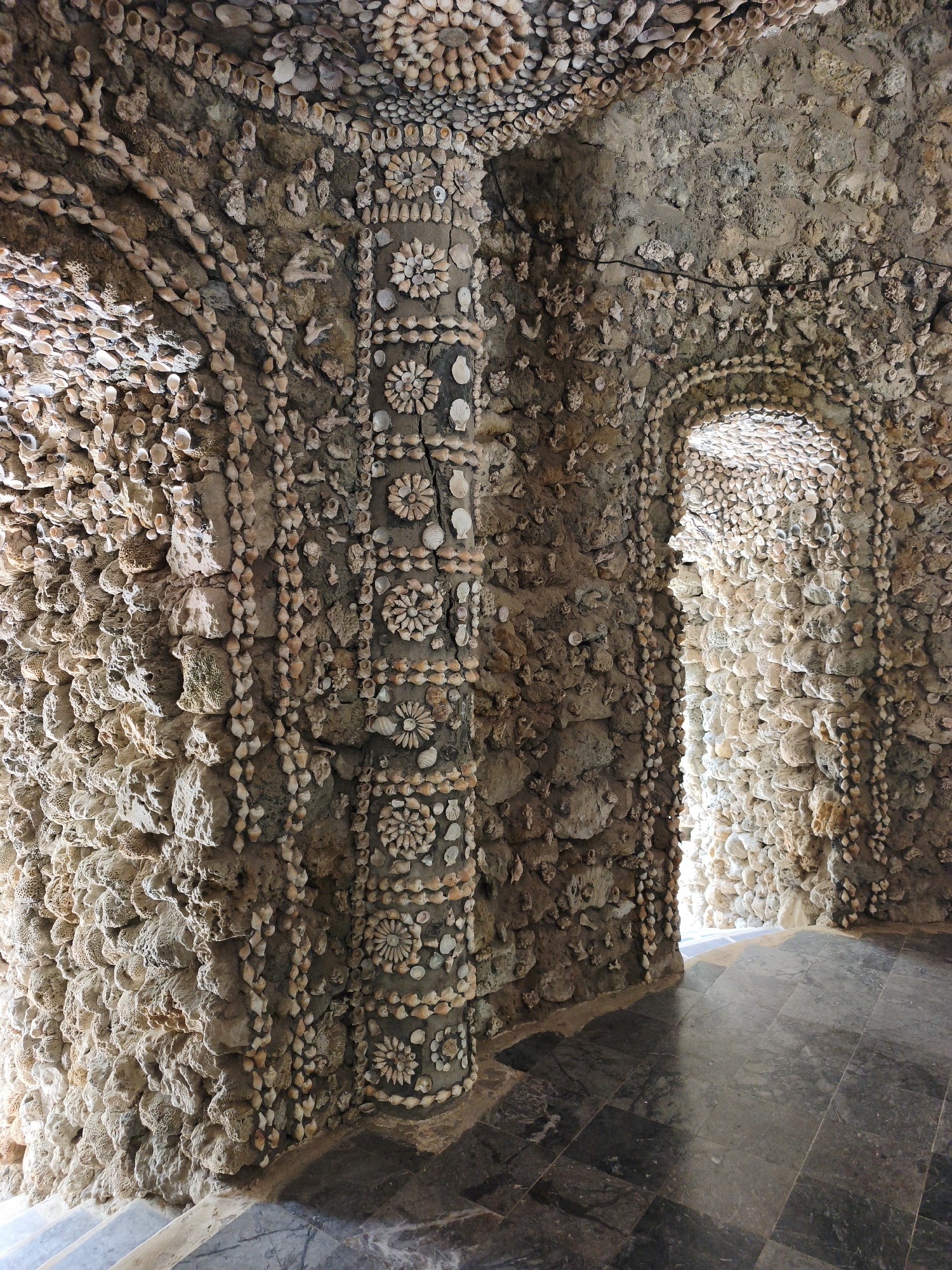The image shows an interior view of a room decorated with shells and rocks. The walls and ceiling are covered in a mosaic of shells and stones, creating intricate patterns. A prominent pillar in the center of the room is also decorated with shells arranged in floral designs. An arched doorway leads to a brightly lit area beyond. The floor is tiled with dark, polished stone. The scene appears to be part of a unique architectural structure, possibly a grotto or a themed building. This type of decoration is reminiscent of the "Van Tuong" or "Nghinh Luong" shell palace in Cam Ranh, Vietnam, known for its extensive use of shells in its construction and ornamentation.