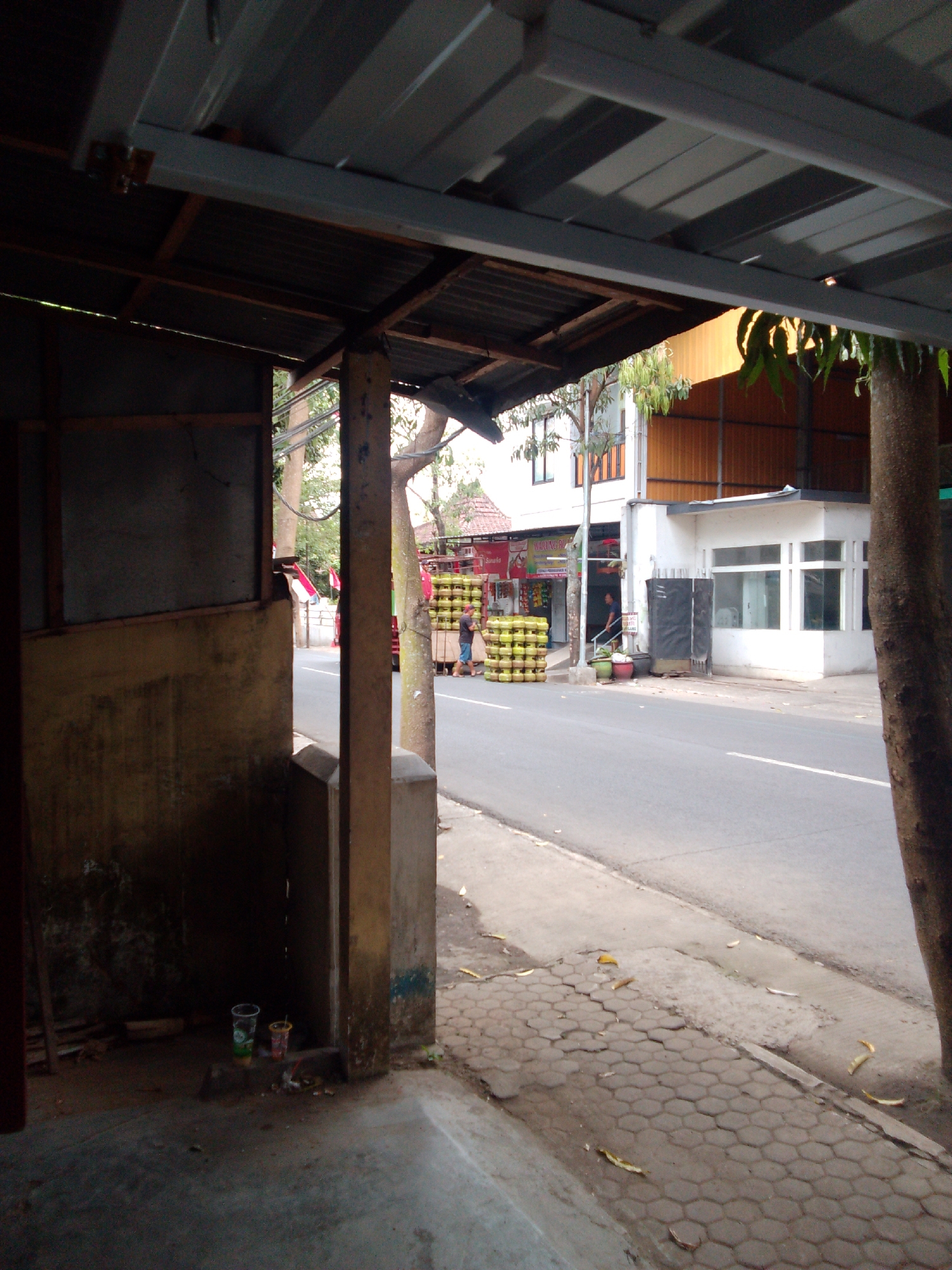 The image captures a street scene in Trenggalek, Indonesia, viewed from a covered outdoor area. The foreground shows a concrete floor and the supports of a roof structure, including a prominent wooden post. To the right, a large tree trunk is visible. The street itself is paved, with a sidewalk in front of it. On the sidewalk and road, stacks of yellow propane tanks are prominently displayed. Two individuals are visible in the mid-ground near the propane tanks, one appearing to be a person in a blue shirt and shorts, and another in a white shirt and dark shorts. Behind the propane tanks, there are buildings, including a white building with large windows and an orange corrugated metal structure. A red truck is also parked near the propane tanks. Red and white Indonesian flags are visible in the background. The overall environment suggests a commercial area, likely a business involved in selling propane.