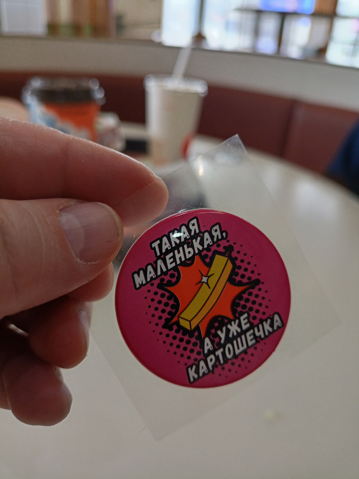 A hand is holding a round sticker with a cartoon illustration of a single french fry with an orange starburst behind it. The sticker has a pink background with black dots. The text on the sticker, written in a sans-serif font, reads "ТАКАЯ МАЛЕНЬКАЯ, А УЖЕ КАРТОШЕЧКА". In the background, there is a table in what appears to be a fast-food restaurant. A cup with a straw is visible in the background. Based on the Russian text on the sticker, the location is likely in Russia. Given that this sticker was reportedly given to employees of a Burger King in Novosibirsk, Russia, it's reasonable to believe that this image was taken there.