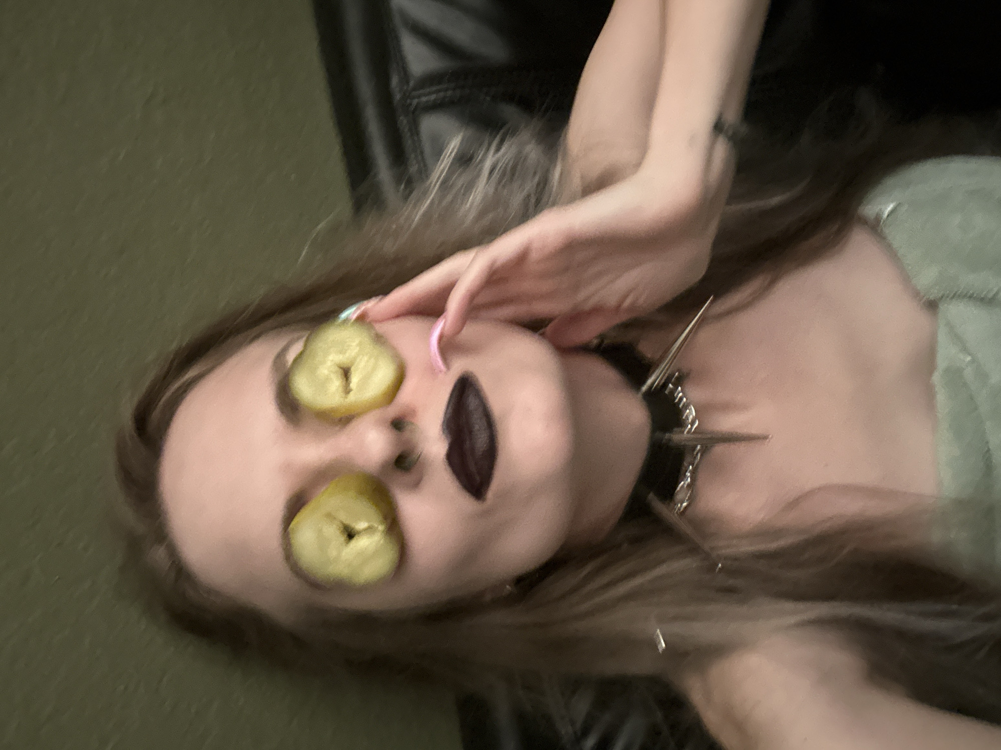 A female is reclining with sliced pickles covering her eyes. Her lips are painted a dark color. She is wearing a spiked collar and a light-colored top. Her left hand is raised towards her face. The background includes a dark-colored surface, possibly a leather chair, and a light green wall. The location is Cape Coral, United States of America.