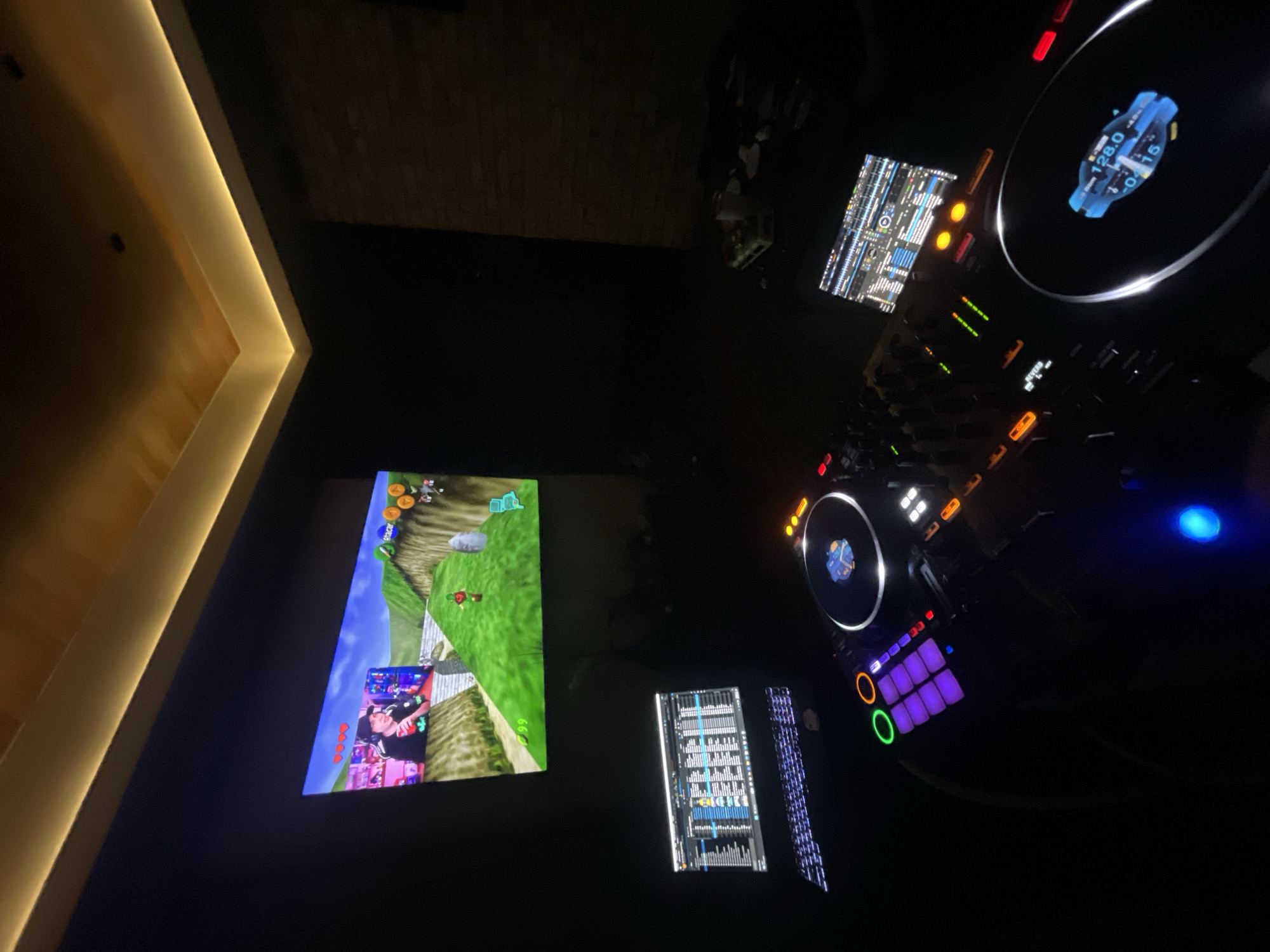 The scene depicts a dark room in Huixquilucan de Degollado, Mexico, primarily illuminated by electronic screens and ceiling cove lighting. In the upper left quadrant, a large screen displays gameplay from "The Legend of Zelda: Ocarina of Time," showing Link in a green, hilly landscape. An overlay on this screen features a male individual wearing a black cap and t-shirt, holding a red can, appearing to be streaming or playing the game. Below the large screen, a professional DJ setup is active. A laptop, displaying DJ software with track lists and waveforms, is positioned behind a DJ controller's left deck. The DJ controller itself, likely a Pioneer DJ model, features illuminated jog wheels (one displaying "128.0" BPM and "-0.15"), glowing faders, knobs, and performance pads in various colors including purple, green, and orange. To the right of the mixer section, another smaller screen or laptop shows additional DJ software interfaces. The room's ceiling features warm, yellow-toned LED strip lighting along its edges. The walls are dark, with a textured surface faintly visible on the right. Some indistinct objects, including a white cup, are present on a surface to the right of the DJ equipment.