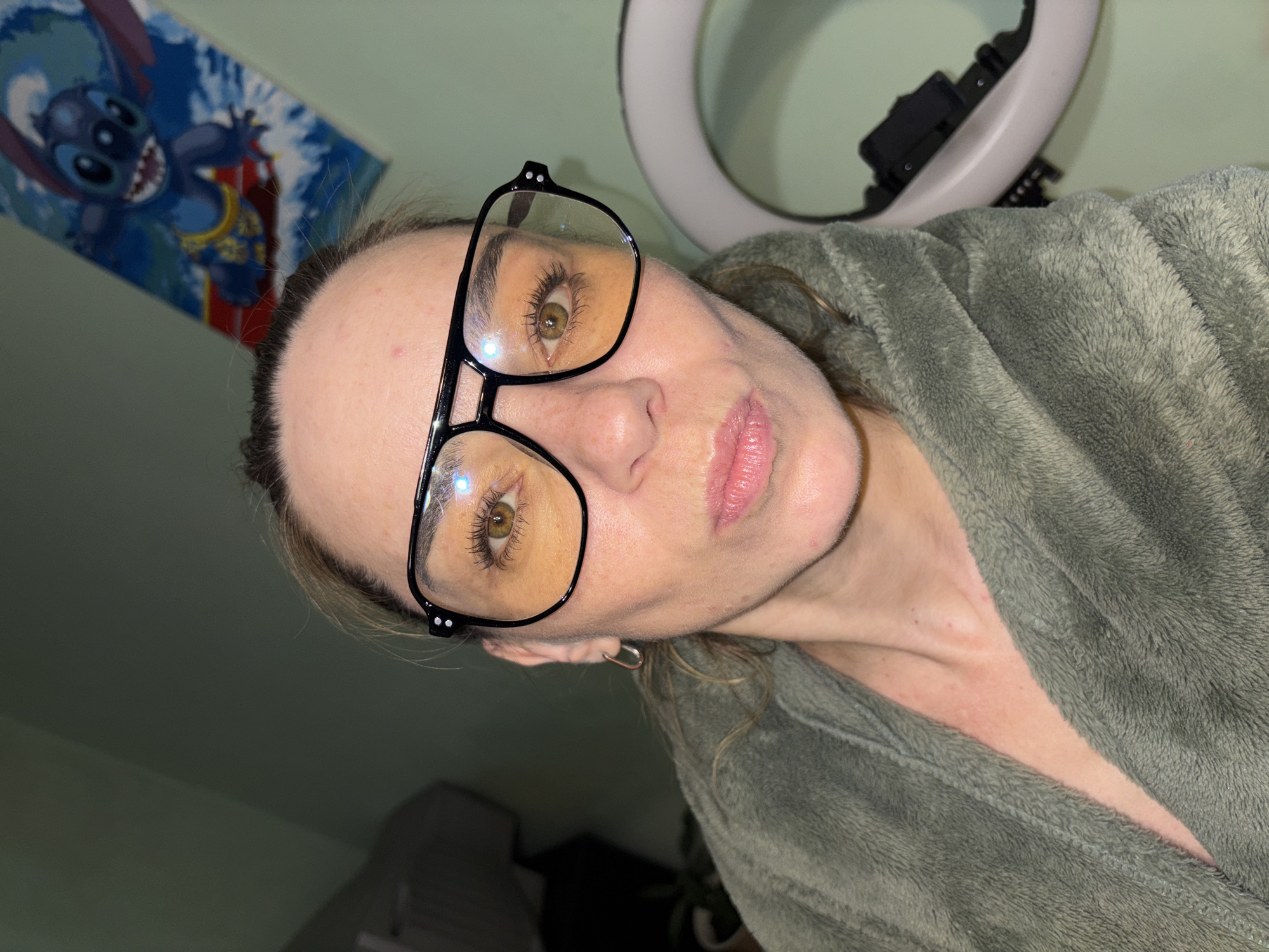 The image shows a close-up, medium shot of a woman's face and upper chest. She is wearing large, black-rimmed glasses with yellow-tinted lenses and a plush, olive-green robe. Her hair is pulled back loosely, and she is looking directly at the camera with a slightly pursed expression. The background is dimly lit, with a light green wall. On the wall behind her, a poster featuring the character Stitch from Disney's Lilo & Stitch is visible. To the right of the woman, a white ring light is partially visible, along with a black electronic device attached to it. The overall lighting suggests an indoor setting, possibly a bedroom or dressing area, at night or in low light conditions. There are no visible indications of the city or specific location.