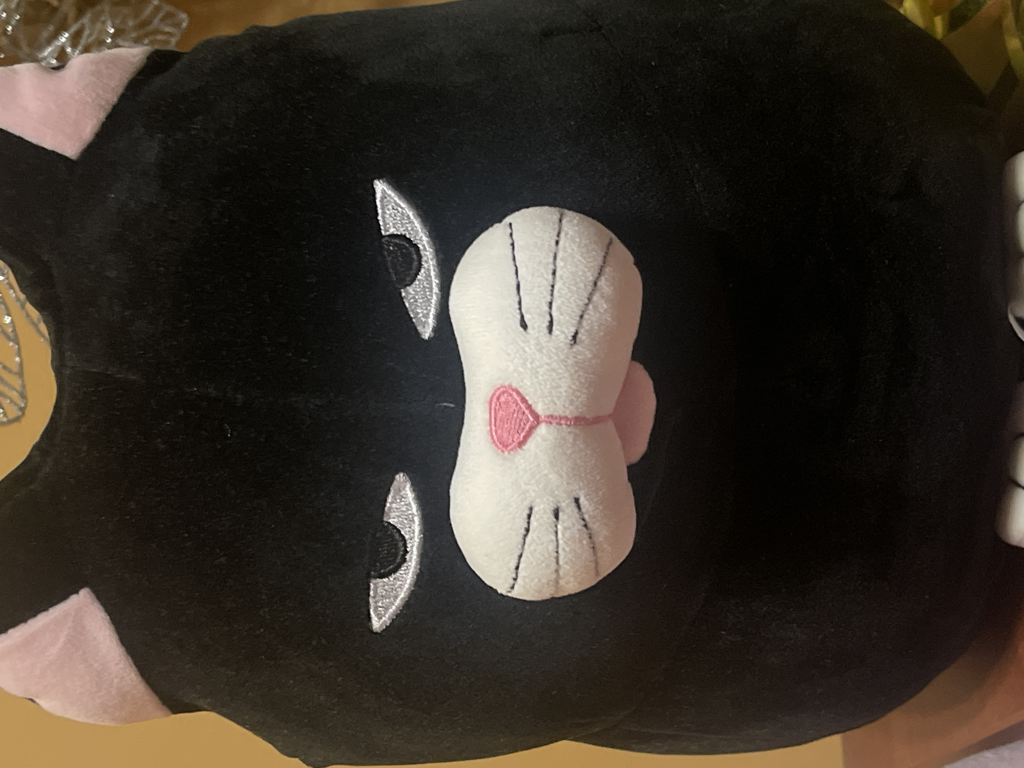 A close-up, straight-on, eye-level shot shows a black plush cat toy with pink ear insides and embroidered facial features. The cat has large, oval, silver embroidered eyes with black pupils, a white muzzle with black embroidered whiskers on either side, and a pink embroidered nose and mouth. The toy appears to be sitting on a surface, possibly a wooden table, with a blurred yellow-orange background. No specific location in Minsk, Belarus, can be identified.