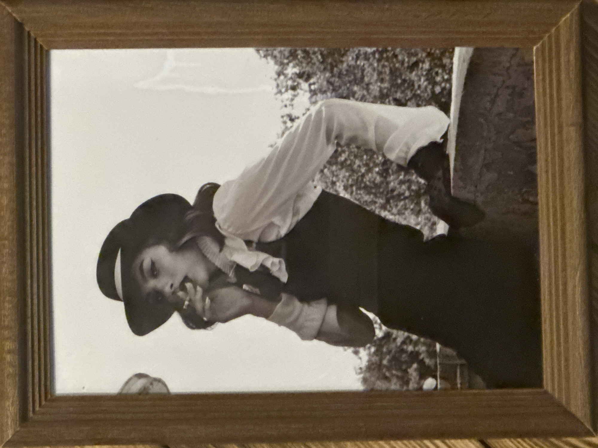 A framed black and white photograph depicts a person in a dark hat, light-colored shirt, and dark vest or dress. They appear to be holding something to their mouth. The background is blurry but suggests trees. The location is unspecified and there is no verifiable information to link it with the city of Selyatino, Russia. The photograph is inside a simple, light-colored wood frame.
