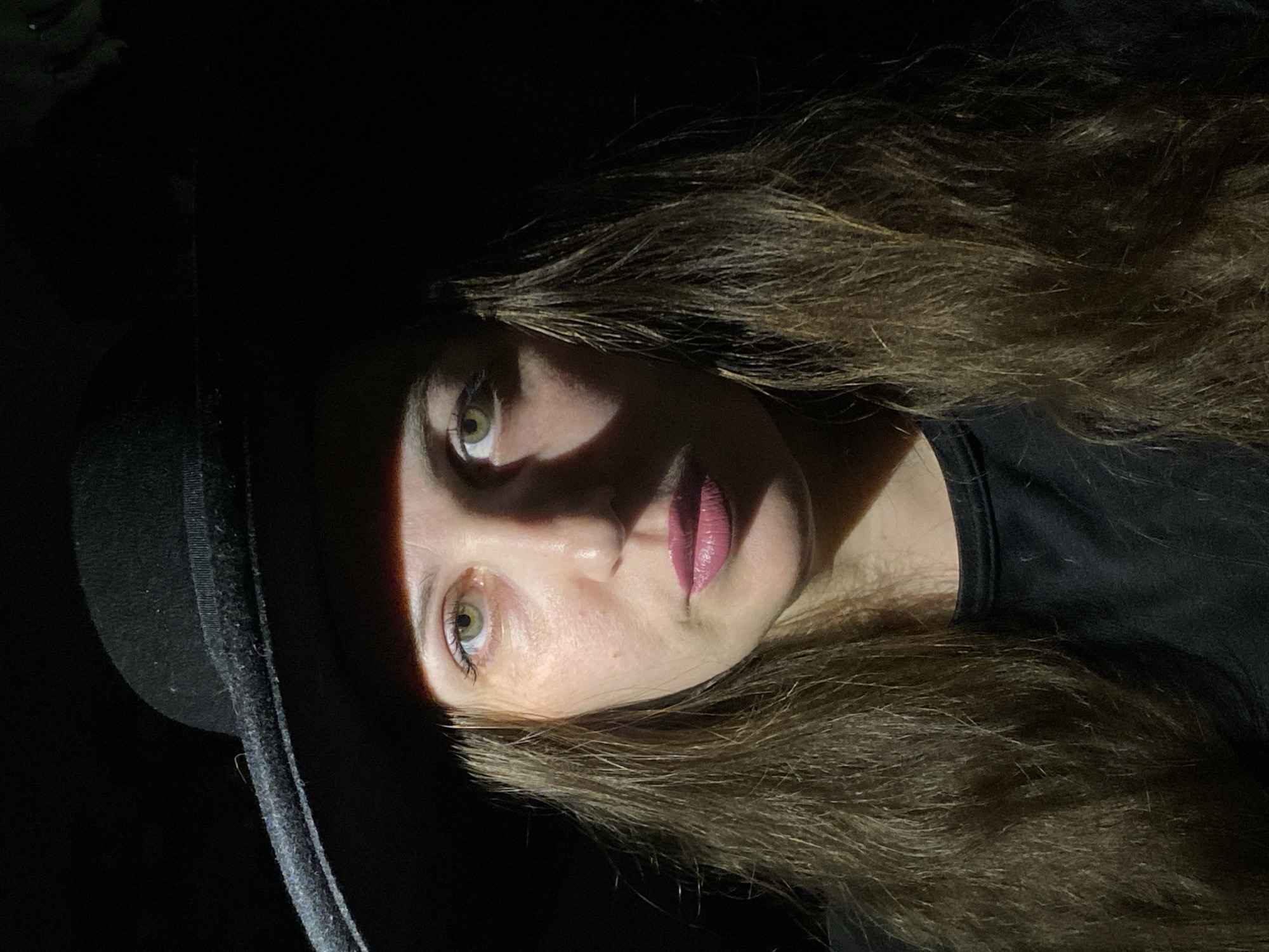 This is a dramatic, close-up portrait of a woman, oriented sideways, depicting her head and shoulders against a completely dark background. The primary subject is a woman with long, wavy, dark brown hair, which cascades around her face and over her shoulders. She wears a black, wide-brimmed hat, visible on the left side of the frame, and a black top or shirt.
Her face is partially illuminated by a strong, narrow beam of light coming from the left, creating stark shadows and highlights. Her left eye (viewer's right) and the left side of her nose and mouth are brightly lit, while the right side of her face is mostly in deep shadow. She has striking green or hazel eyes, with defined eyelashes, and her lips are painted with a soft pink or mauve lipstick. Her gaze is directed slightly upwards and towards the viewer, conveying a thoughtful or intense expression.
The setting appears to be dark, likely at night or in a dimly lit indoor space, emphasizing the dramatic lighting effect. There are no discernible details in the background other than absolute darkness, suggesting a controlled photographic environment. The activity is a posed portrait, utilizing high contrast lighting to create a mysterious and artistic mood. No text is visible in the image. The location context provided is Markovskiy, Russia.