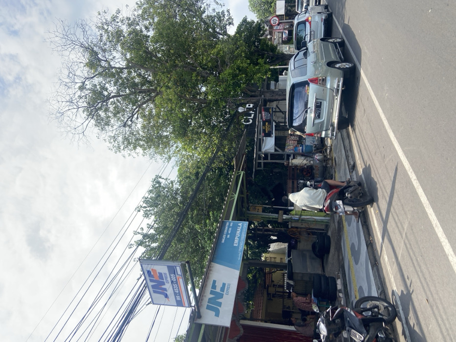 The image shows a street in Mojokerto, Indonesia, located at coordinates -7.467 latitude and 112.445 longitude. The street is lined with parked cars, including a silver minivan, a white minivan, and a white sedan. A man in a white hooded sweatshirt is riding a black motorcycle in the street. There is a JNE Express sign on the front of a building to the left of the street. The road surface is paved, and there are white lines marking the lanes. The road is bordered by a sidewalk with a grey curb. The image was taken during the day, with a bright blue sky and some cloud cover. There are trees lining the street. The image is framed from a perspective of a pedestrian standing on the sidewalk to the left of the street. The image is taken from a low angle.