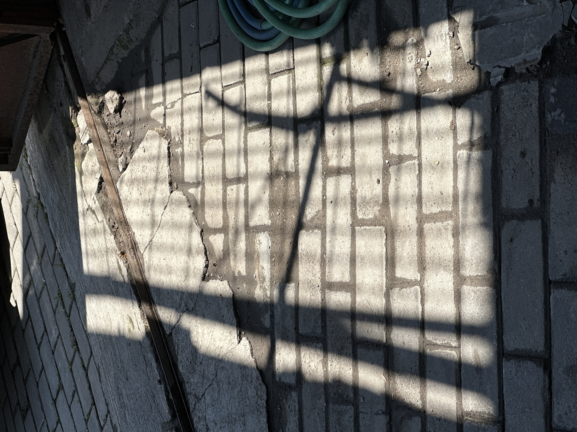 In the city of Bulakrejo, Indonesia, an outdoor scene features a sun-dappled brick pathway with a visible shadow pattern cast from what appears to be a fence or grate. The pathway, composed of rectangular bricks laid in a linear pattern, shows signs of wear and tear, with some cracks and areas of loose dirt and small stones. To the right of the pathway, a green and blue garden hose is coiled. The lighting suggests it is daytime, with strong sunlight creating the distinct shadows.