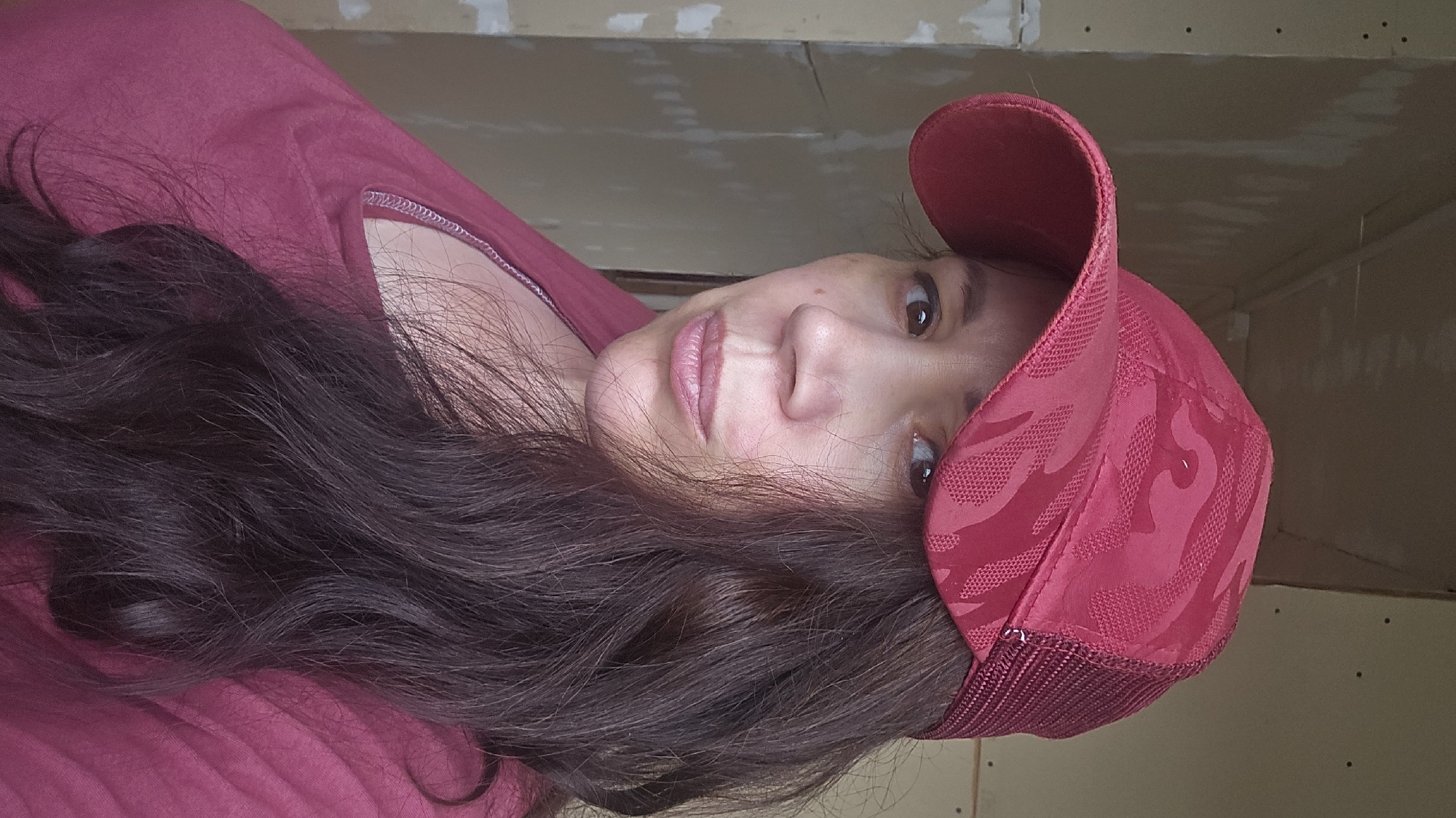 A medium shot shows a woman from the shoulders up, looking slightly off-camera to the left. She is wearing a dark red baseball cap with a camouflage pattern and a mesh back, and a matching dark red t-shirt. Her long, dark brown hair is visible on the right side of the frame. The background appears to be an unfinished interior space with light-colored walls that have exposed studs and visible markings. The lighting suggests an indoor environment, likely during the day. There are no specific landmarks or details to place this image within the city of Ермолаево, Russia.