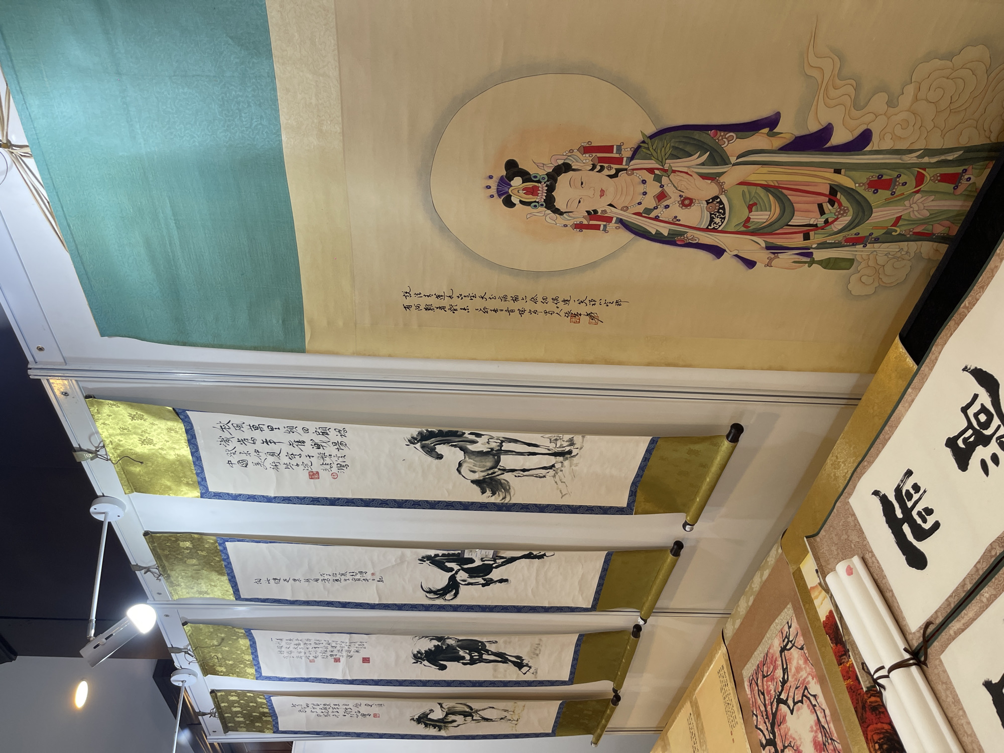 The image shows a display of traditional Chinese art, likely at an exhibition or market stall. The focal point is a large scroll painting of a female deity, possibly Guanyin, depicted with a serene expression and elaborate adornments, standing amidst swirling clouds. To the left of this central piece are four vertical scrolls, each featuring a ink painting of a horse in a different pose. These scrolls are adorned with golden fabric at the top and blue patterned borders. Below these, on a table, are several rolled-up scrolls and a framed artwork depicting a flowering tree with pink blossoms. The background appears to be a white partition wall. The overall scene suggests a cultural exhibition of art from China, possibly in Futian District, known for its art markets and galleries.