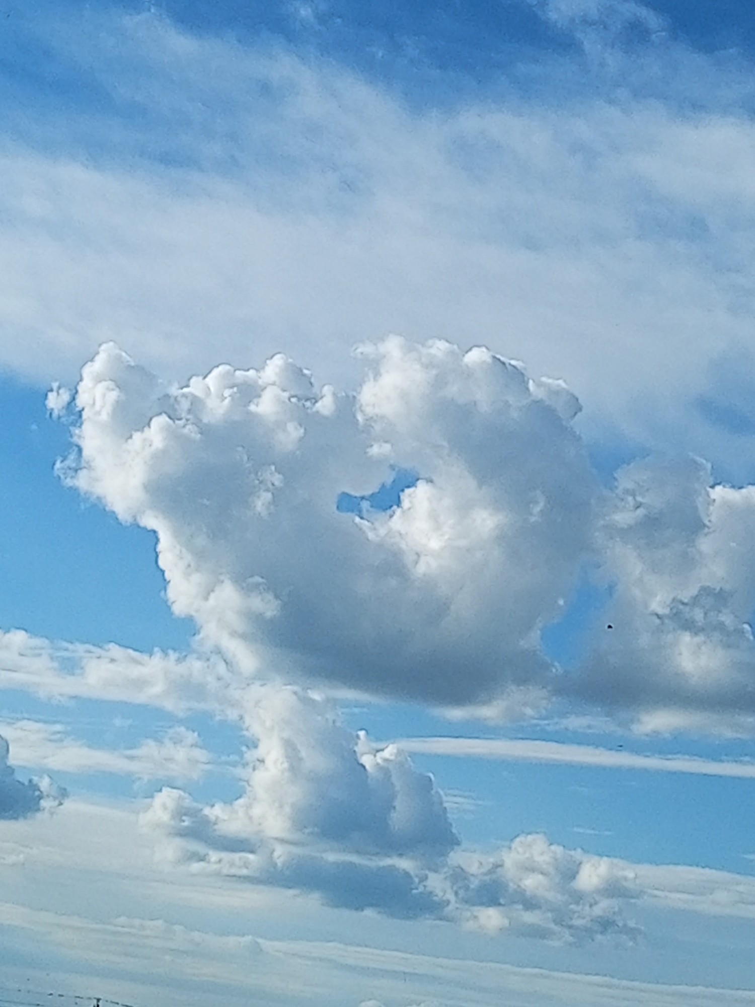 A photograph captures a bright blue sky filled with various types of clouds. The dominant cloud formation in the upper center is a large, puffy cumulus cloud with a distinct heart-shaped opening in its center, revealing the blue sky behind it. To the left, a smaller cluster of cumulus clouds appears to be detached from the main formation. In the lower part of the image, several layers of thinner, wispy clouds are visible, creating a sense of depth. The overall lighting suggests it is daytime, possibly late morning or early afternoon, given the brightness of the sky and the shadows within the clouds. There is no discernible indication of the city of Бөрлі, Kazakhstan within the frame; the image solely focuses on the sky and clouds. No people, subjects, objects, actions, or interactions are present in the image. The environment is purely atmospheric.