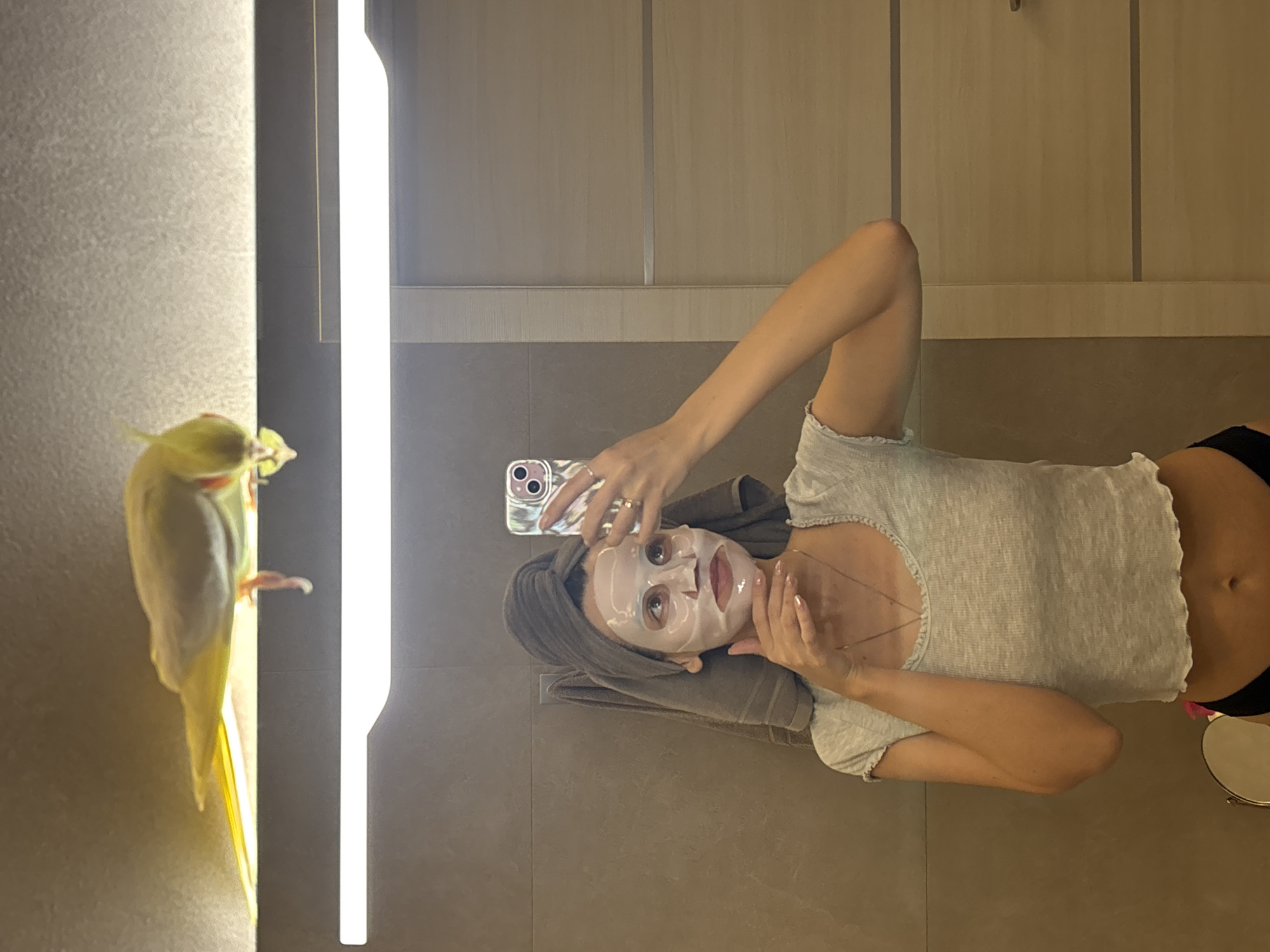 The scene depicts a bathroom interior in a private residence in Krasnoyarsk, Russia. A female subject is captured in a mirror selfie, extending from her mid-torso upwards. She is wearing a white sheet mask on her face and has her hair wrapped in a grey towel. Her attire consists of a light grey, ribbed, short-sleeved crop top and a visible portion of black undergarment. A thin necklace and a ring on her left ring finger are present. She holds a smartphone with a distinctive case in her right hand to take the photograph, while her left hand is positioned beneath her chin. The background features light brown tiled walls, a wooden-paneled door or cabinet, and a grey towel hanging on a hook. Above a prominent linear light fixture, a light yellow-green bird, appearing to be a parrot, is perched, looking downwards towards the reflection. A small, circular magnifying mirror is partially visible in the lower-left corner of the frame.
