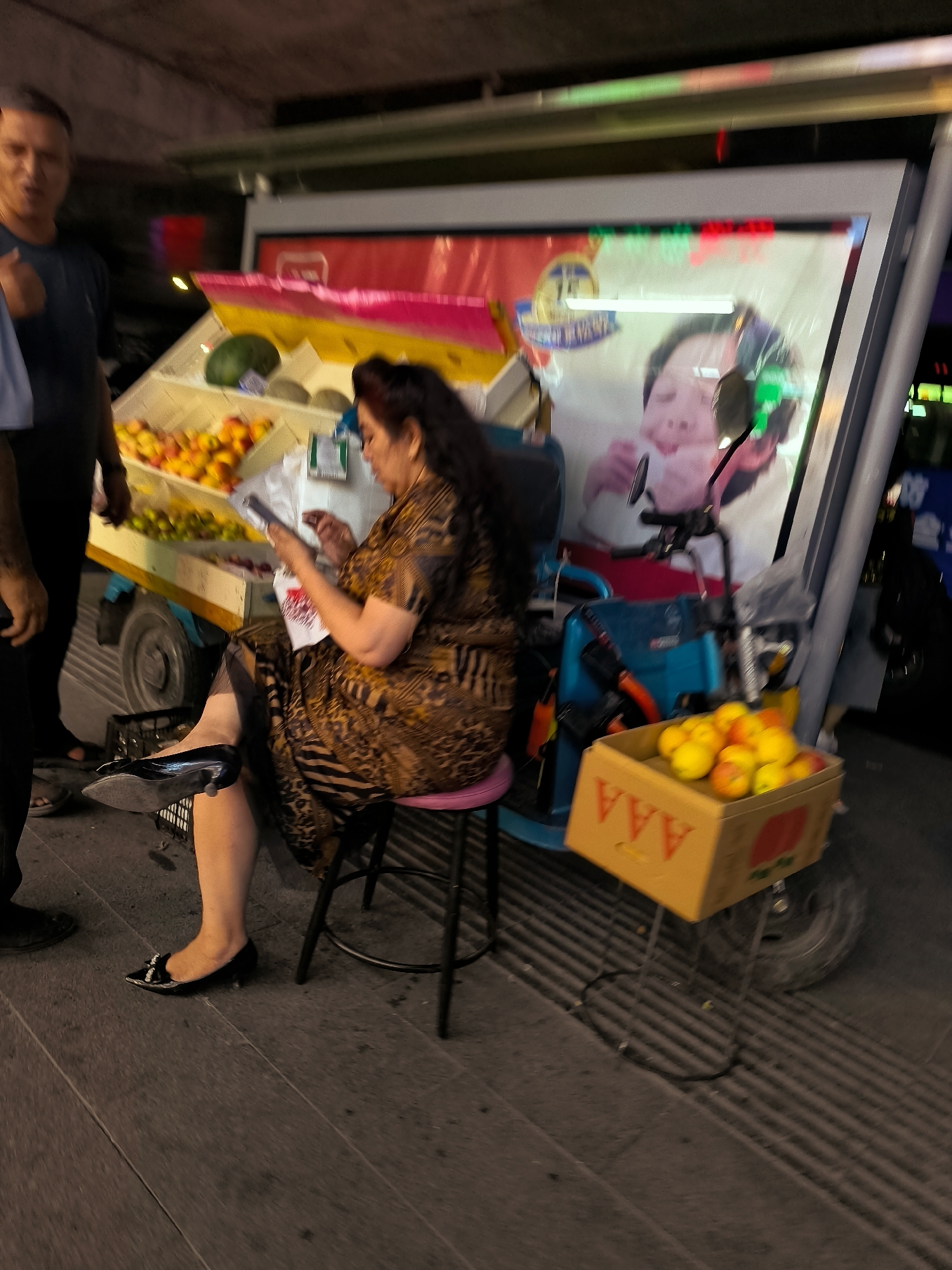 A medium shot captures a busy street scene at dusk in Ürümqi, China, featuring a woman selling fruit from a mobile cart. The woman, seated on a pink stool, is dressed in a patterned brown dress and black high-heeled shoes. She is focused on her mobile phone, which she holds in her hands. The fruit cart, a blue and silver structure, displays various fruits in tiered baskets. A watermelon is visible in the upper left basket. In the foreground, a yellow box contains a pile of oranges. To the left of the cart, a man in a dark t-shirt stands, partially visible, with his hand raised. The background is blurred, suggesting a bustling urban environment with neon lights and other vehicles. The overall impression is one of everyday commerce taking place in a lively city setting.
