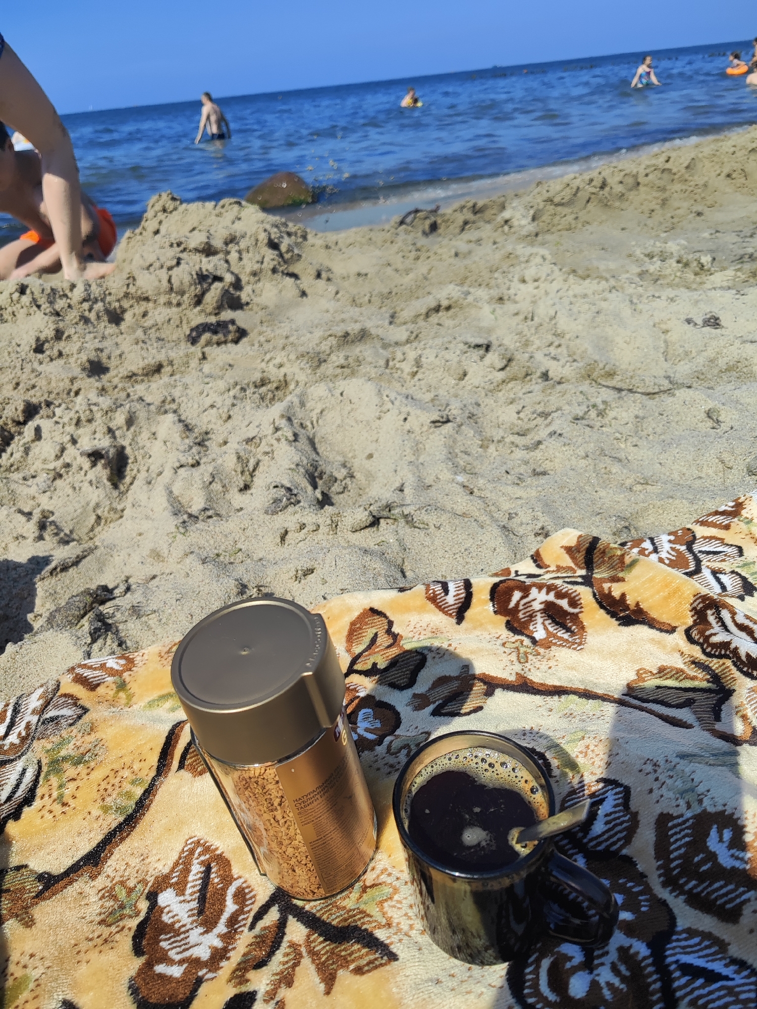 The image shows a beach scene in Pionersky, Russia, with the Baltic Sea in the background. In the foreground, a beach towel with a floral pattern is spread on the sand. On the towel are a glass jar of instant coffee granules and a black mug filled with black coffee, with a spoon in it. The sea is blue and calm, with a few people swimming or wading in the water. The sand is light brown and appears dry. The sky is clear and blue, indicating a sunny day.
