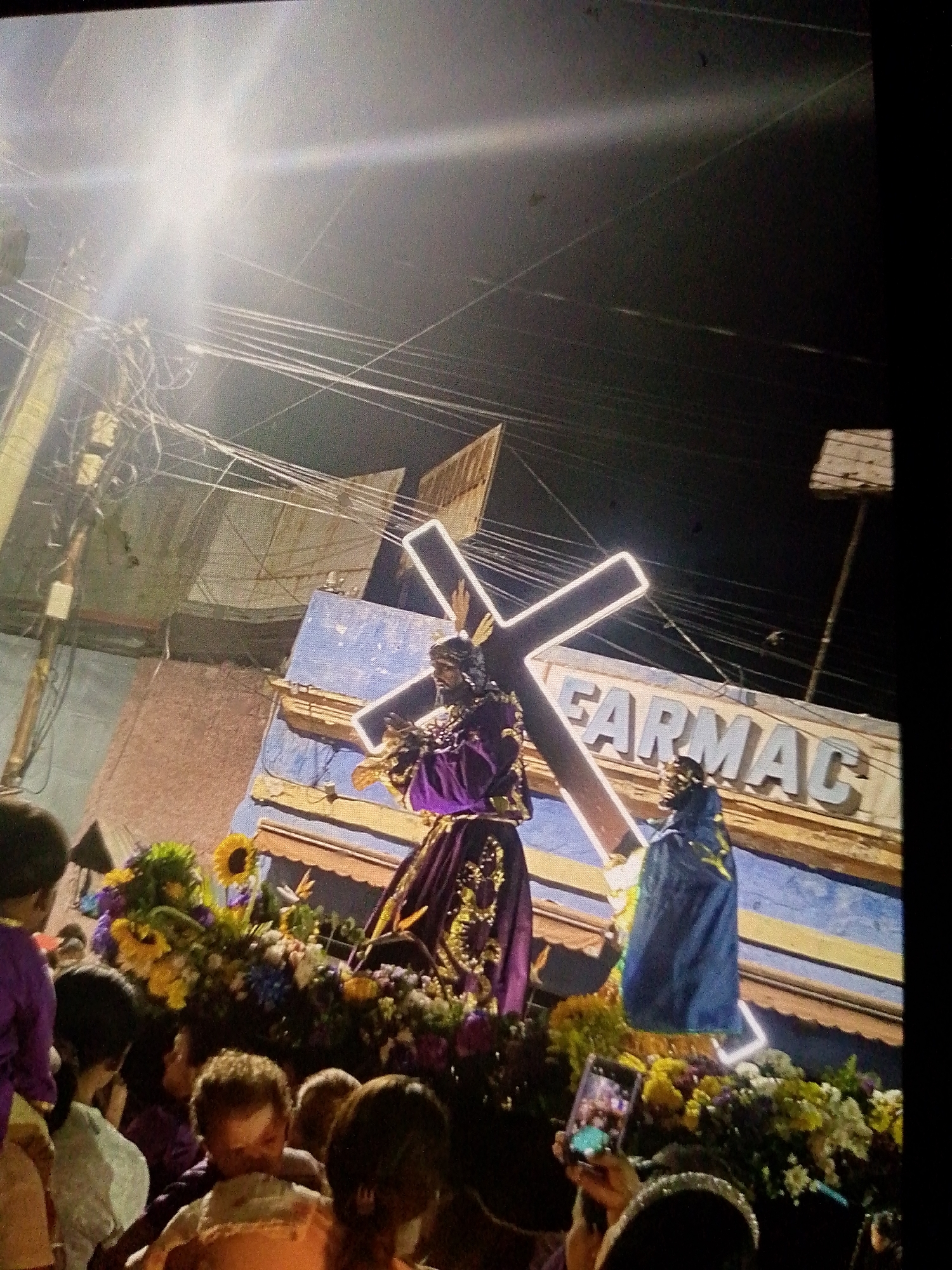The image shows a nighttime religious procession in Caracas, Venezuela. A statue of Jesus carrying the cross, illuminated with glowing lights, is the central figure. The statue is adorned in purple and gold robes and surrounded by flowers, including sunflowers. Another figure in a blue robe stands beside it. A crowd of people, mostly facing the statue, are observing the procession. Some are taking photos with their phones. The background includes a building with the word "FARMAC" visible on it. Overhead, there are numerous wires and a bright light source. The overall atmosphere suggests a solemn religious event taking place in an urban setting.