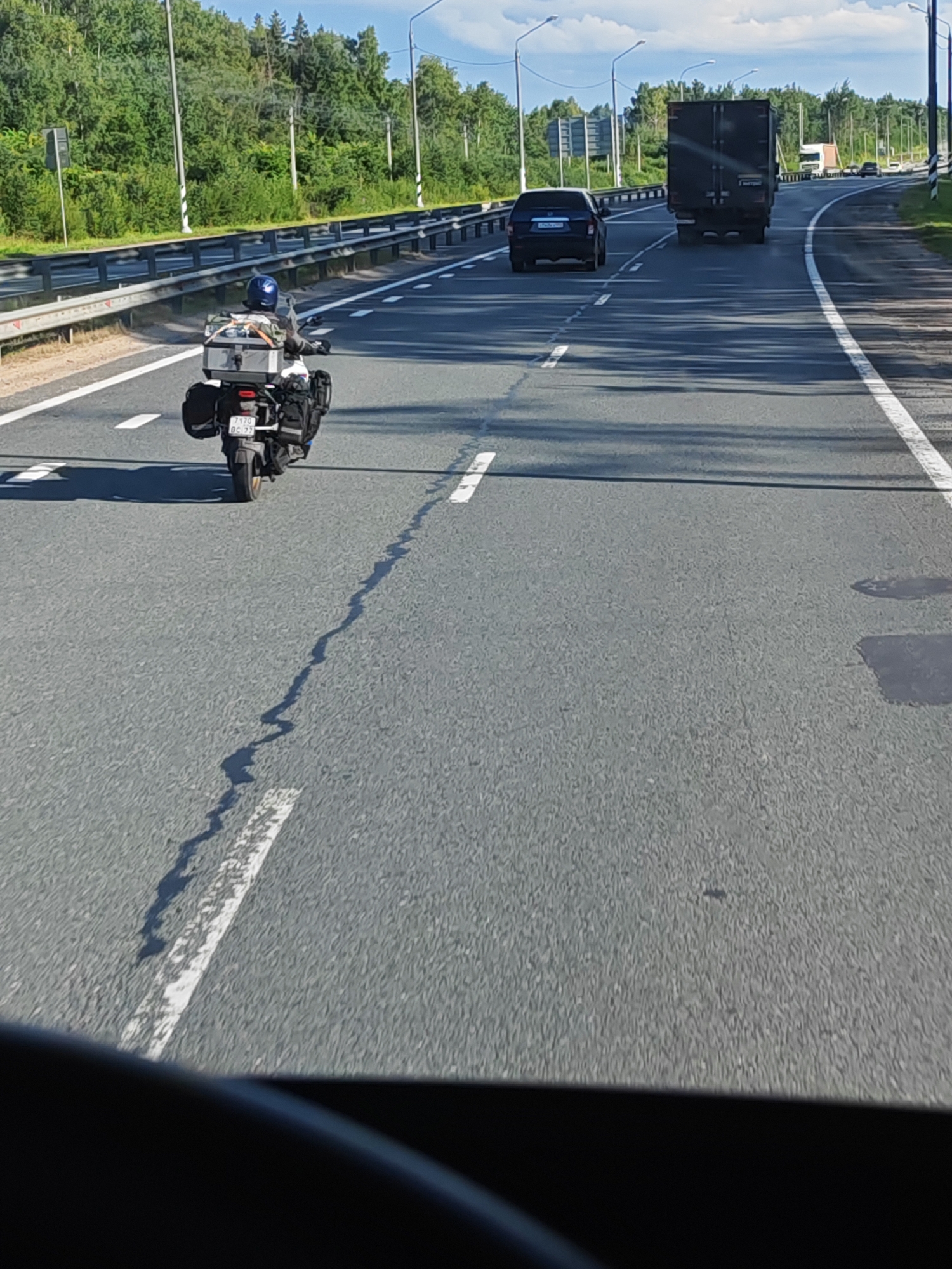 A motorcyclist wearing a blue helmet is riding on a highway in Torzhok, Russia. The motorcycle is equipped with luggage racks on the back and sides, carrying a silver case and black bags. The motorcycle's license plate reads "7170 BC 77". The road has white lane markings and guardrails are visible on the left side. Other vehicles, including a black car and a large dark truck, are also present on the highway. The surrounding environment is characterized by green trees and a blue sky with some clouds. The image appears to be taken from a vehicle following the motorcycle.