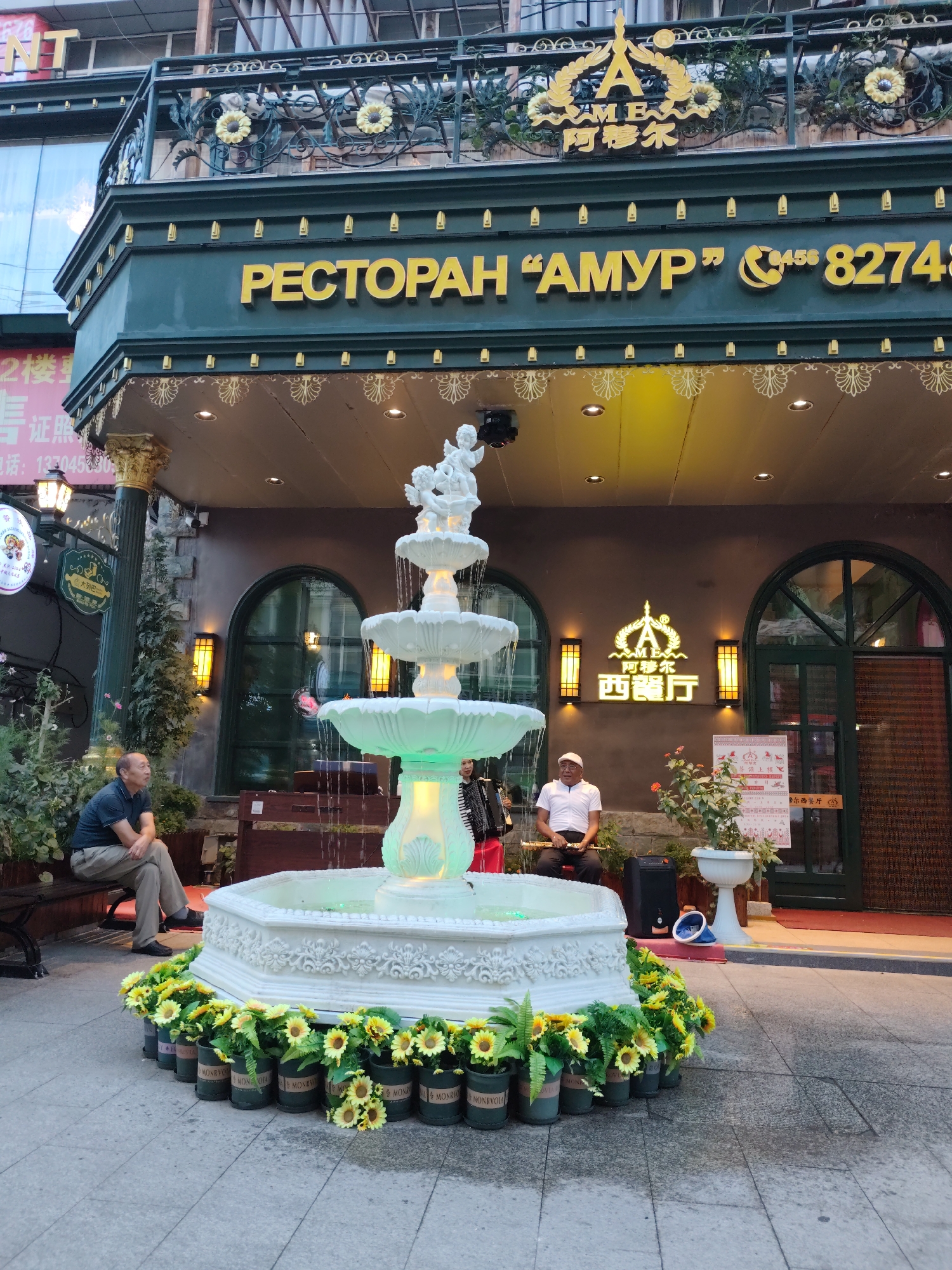 In the city of Aihui, China, a tiered fountain with white sculpted cherubs on top is illuminated by green light, with water cascading down its levels. Surrounding the base of the fountain are numerous potted sunflowers. To the left, a man in a dark polo shirt and light-colored pants sits on a dark bench, looking towards the fountain. To the right of the fountain, a man in a white polo shirt and white cap sits on a bench, holding a flute-like instrument. Behind him, a woman plays an accordion. The fountain is situated in front of a building with a green facade, featuring large yellow letters spelling "РЕСТОРАН “АМУР”" and a phone number. Above the entrance, a golden crest with the letter "A" and Chinese characters is visible. The scene appears to be outdoors, possibly a pedestrian area or a plaza, with a tiled ground.