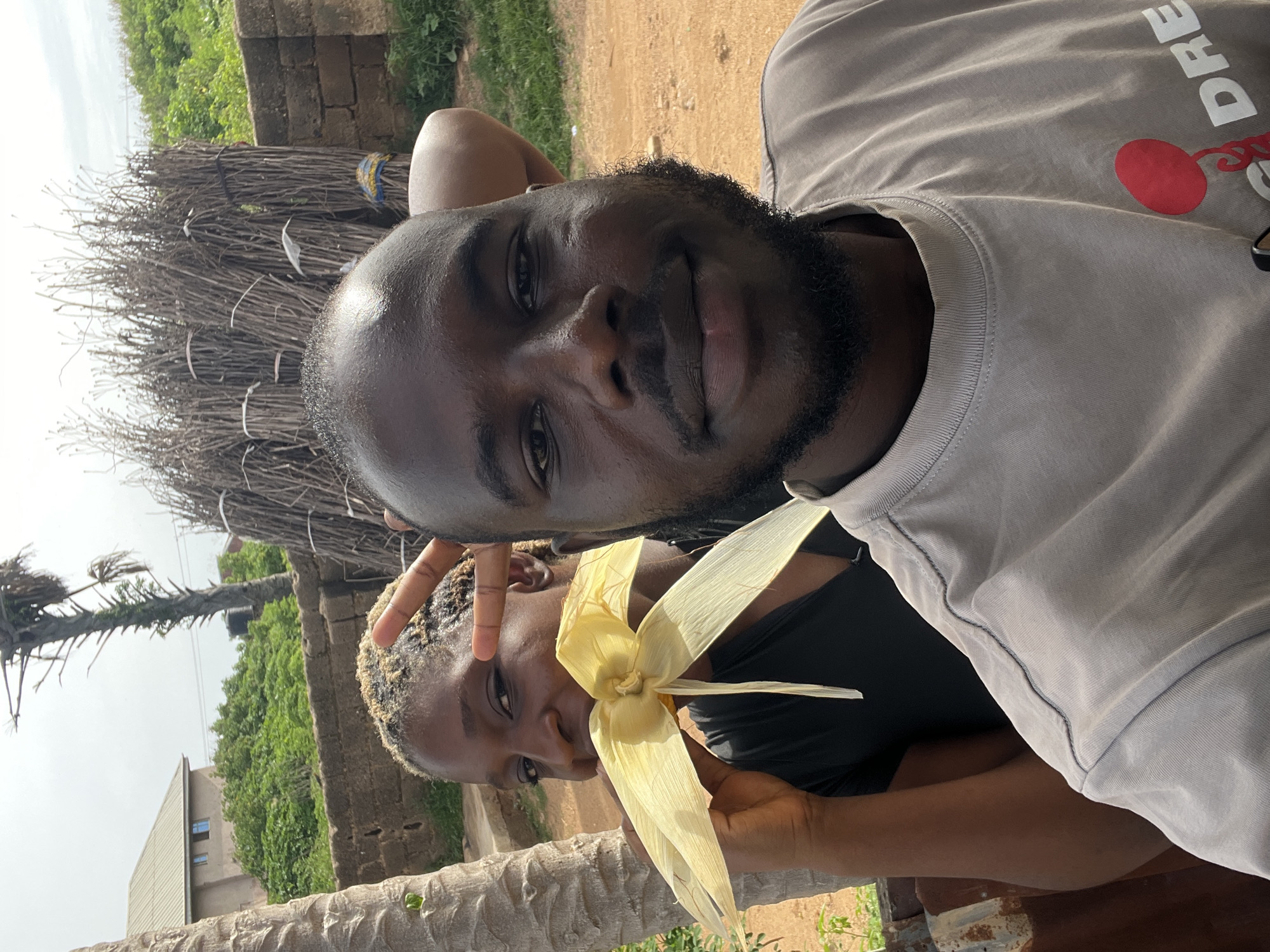 The image shows two people, a man and a woman, outdoors in what appears to be a rural or semi-rural setting in Jos, Nigeria. The man, in the foreground, is wearing a light gray t-shirt with text that is partially visible. He has dark skin and a beard. He is looking towards the camera with a slight smile. The woman is to his left, and she is holding what looks like corn husks to her face, possibly for a decoration or a game. She has dark skin and is wearing a black top.
Behind the people, there is a large bundle of sticks or branches tied together with white and blue string. Further in the background, there is lush green vegetation, a brick wall, and part of a building. The lighting suggests it is daytime, and the sky is overcast. The overall impression is one of a casual, everyday moment captured in a natural environment. The image seems to be a selfie, as indicated by the man's pose and the framing of the shot. The woman's expression is playful.