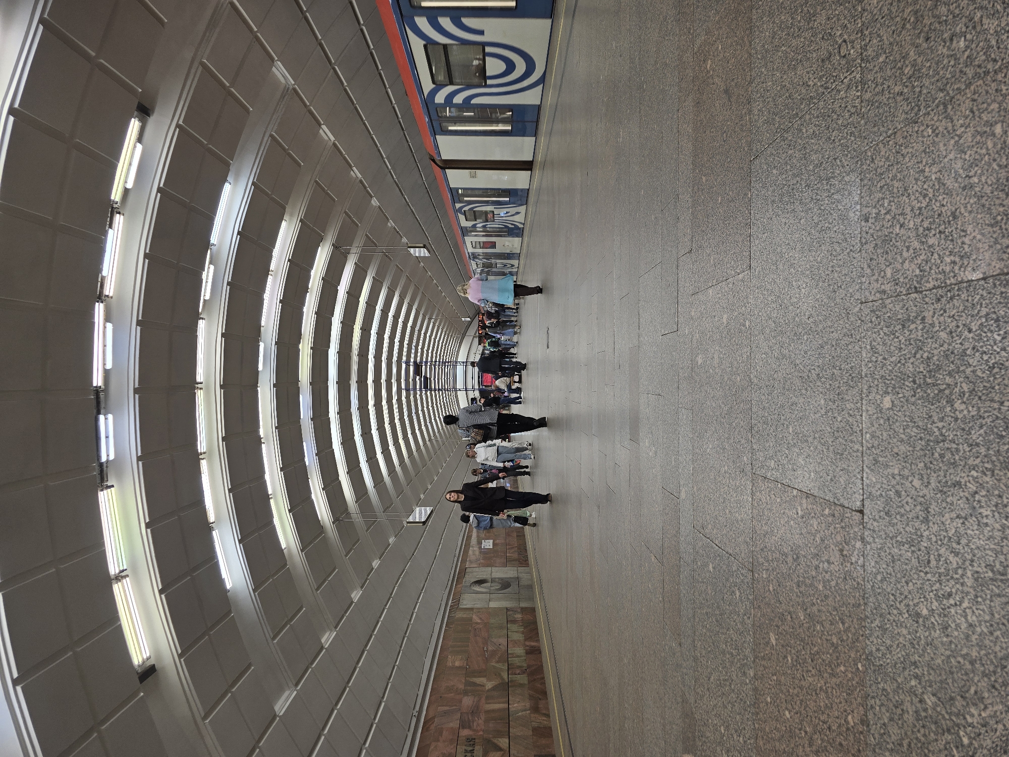 The image depicts the interior of a subway station. A subway train is visible on the right side of the frame. A platform made of grey, granite-like tiles stretches from the bottom to the center of the image. Several people are standing or walking along the platform. The left side of the image features a curved wall with rows of fluorescent lights. At the bottom left of the frame, there's a section of patterned, marble-like wall beneath the curved wall with the lights. The scene conveys a sense of urban transit and public space.