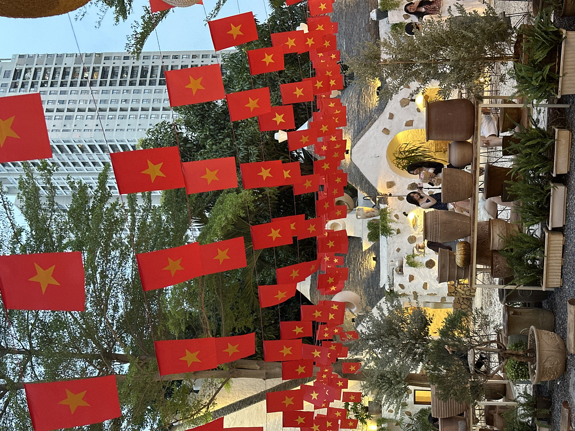 The image shows a street decorated with numerous red Vietnamese flags, each featuring a yellow star. These flags are strung overhead, creating a festive and patriotic atmosphere. In the background, a modern high-rise building stands, contrasting with the more traditional decorations.
On the right side of the image, there is a building or establishment with a white facade, decorated with stones and a decorative archway. People are sitting at tables within this establishment. The location is Thủ Đức, Vietnam.
Plants and decorative pots adorn the sidewalk, contributing to the overall ambiance. The image is taken at an angle, looking up at the flags and towards the building.
