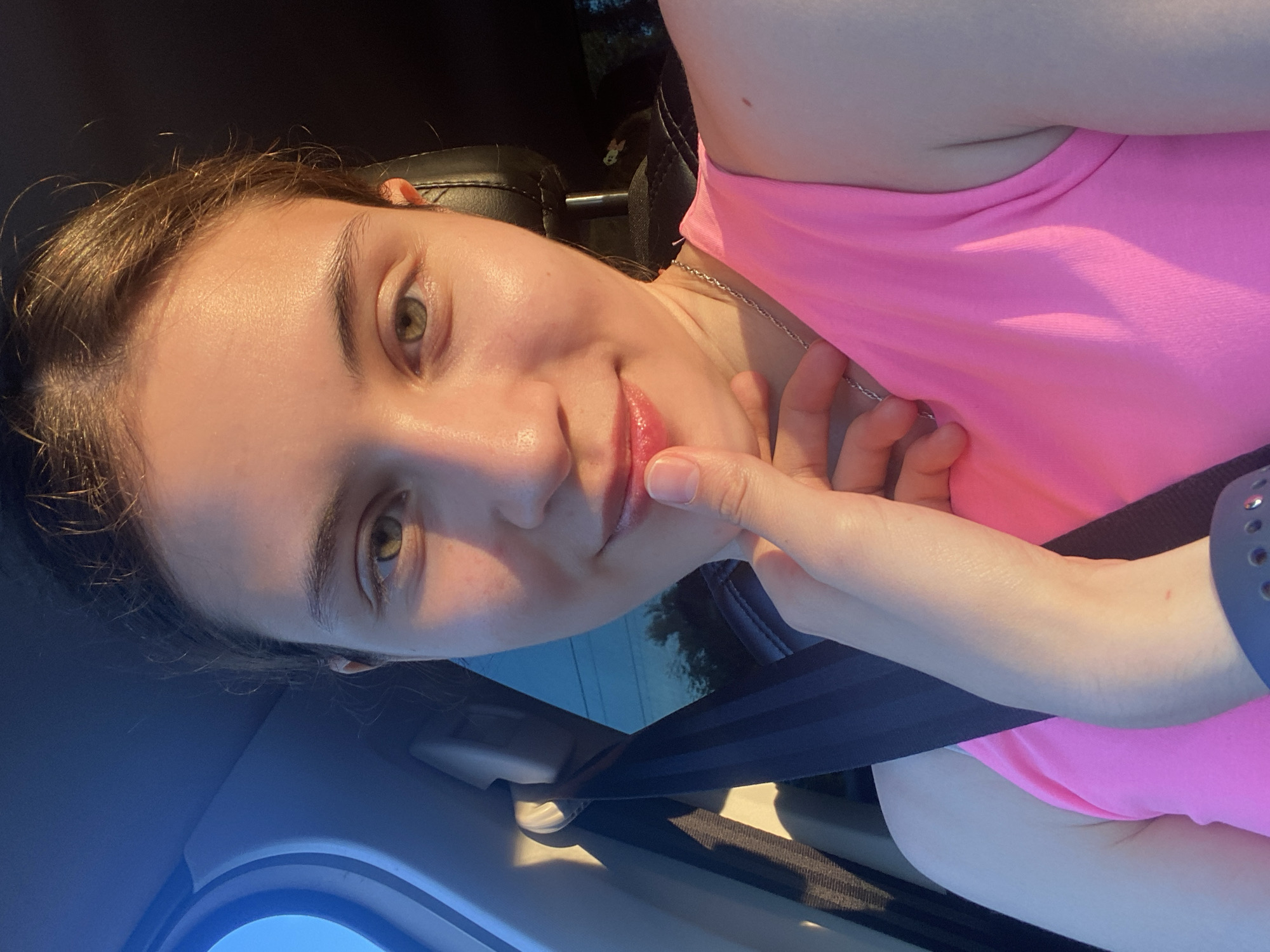The image shows a young woman sitting in the passenger seat of a car, likely on a sunny day. She is wearing a pink tank top and a silver necklace. Her hand is gently touching her lips, and she is looking towards the camera with a slight smile. The interior of the car is visible, including a black seatbelt and the edge of a car window. The lighting suggests that the sun is setting or rising, casting a warm glow on her face. The background outside the window is blurred, showing a hint of trees and power lines, suggesting an urban or suburban setting in Votkinsk, Russia.