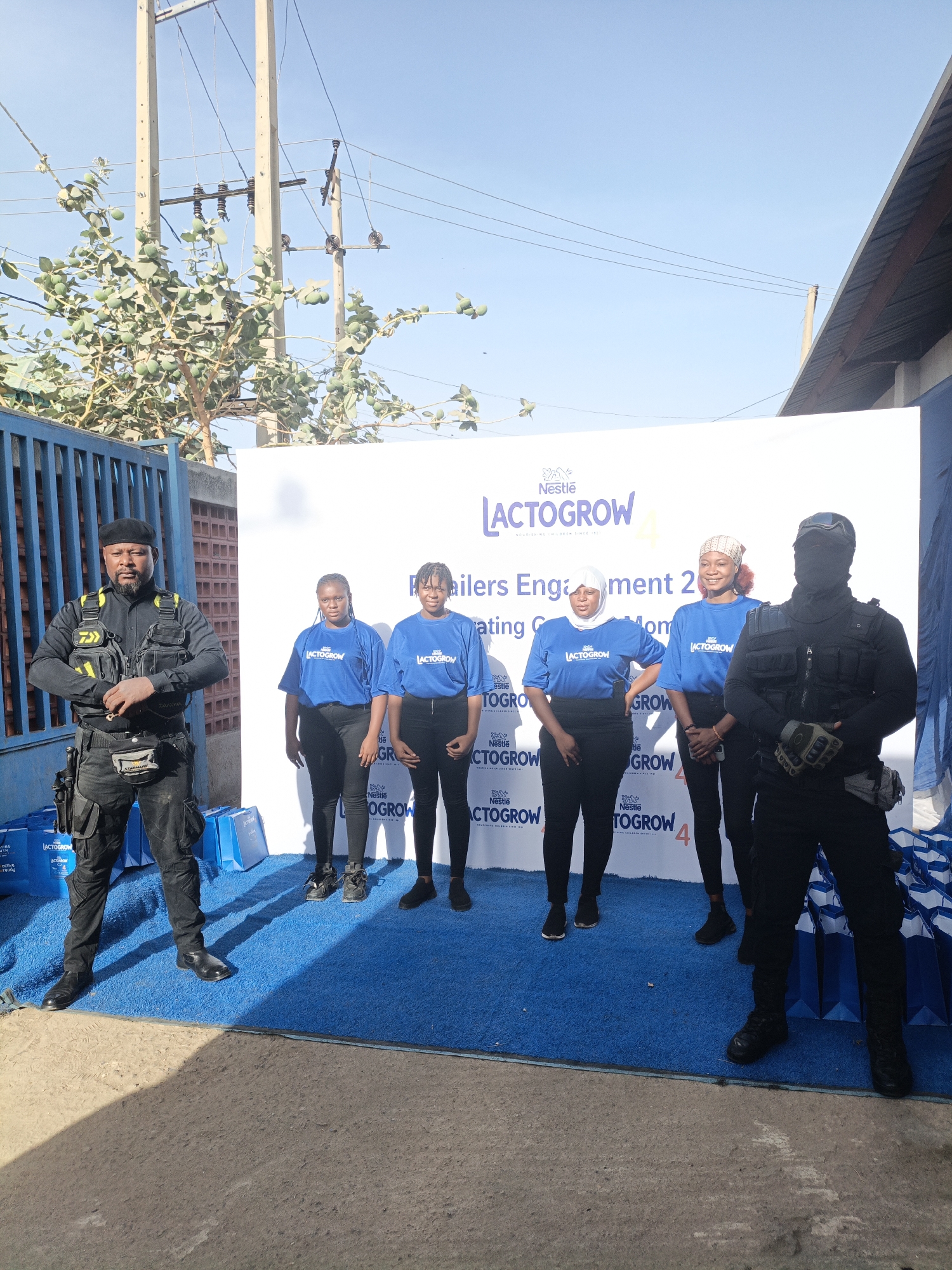 The image captures an outdoor promotional event likely sponsored by Nestlé Lactogrow, in what appears to be Tarauni, Nigeria. At the center, five women stand in front of a white backdrop displaying the Nestlé Lactogrow logo and the text "Retailers Engagement 20," followed by the words "Creating G Mom." Each woman wears a blue T-shirt with the Lactogrow logo and black trousers.
To the left and right stand two security guards dressed in full tactical gear, including vests, belts with pouches, and headwear. The guard on the right has their face covered. The setting includes a blue carpet under the subjects and branded promotional materials in the background. The surroundings suggest an urban environment with power lines and buildings visible in the background. The lighting indicates it is daytime.