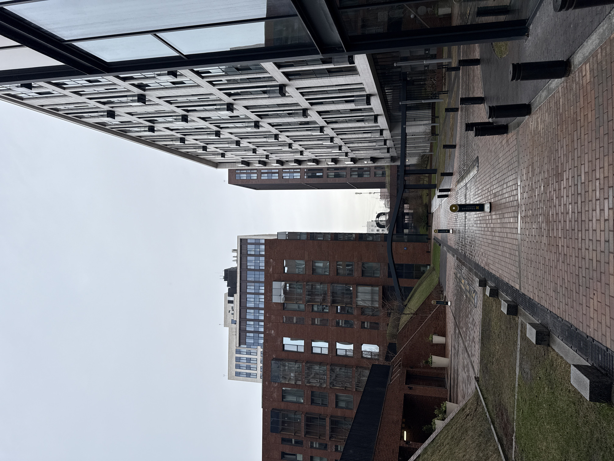 The image depicts a street scene in Moscow, Russia, featuring several modern buildings with distinctive architectural designs. The buildings are primarily constructed with brick and glass, with a mix of low-rise and high-rise structures. The street is paved with red brick and includes decorative elements such as short, black bollards and sections of landscaping with grass. The sky is overcast, suggesting a grey or rainy day. A small, black signpost with yellow lettering, likely indicating a street name or direction, is visible on the street. There are no people present in the immediate view.