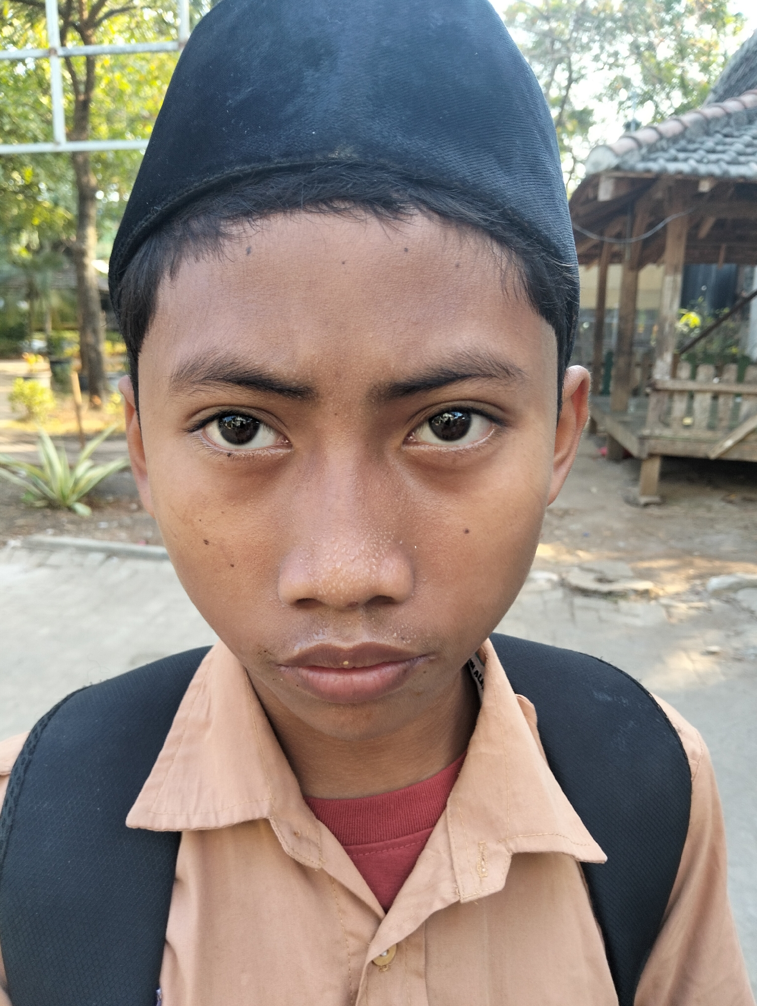 A close-up, eye-level, portrait-style photograph features a young boy wearing a black cap and a tan collared shirt. His gaze is directed straight at the camera, with his mouth slightly open. A black backpack is visible over his shoulders. The background is blurred, showing trees, a wooden structure with a tiled roof, and a paved surface, suggestive of an outdoor, possibly school or village, setting in Paciran, Indonesia. The lighting indicates it is daytime, possibly late afternoon due to the warm tones and shadows.