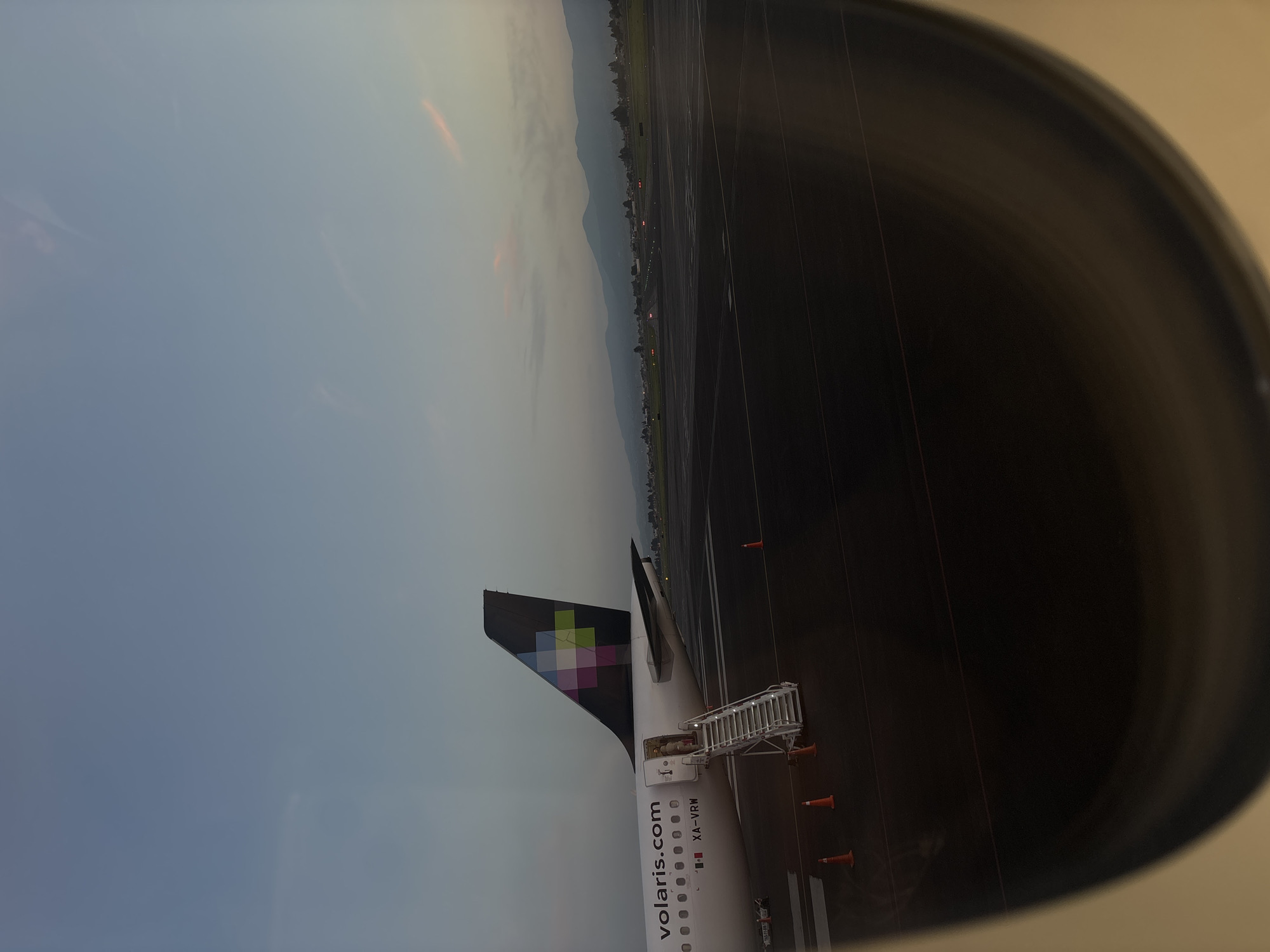 The image is taken from the window of an airplane, showing part of the wing and tail of a Volaris airplane. A set of stairs is connected to the plane's door. There are orange traffic cones on the tarmac surrounding the plane. In the background, a runway and some buildings can be seen, and further in the distance are mountains. The sky is overcast.