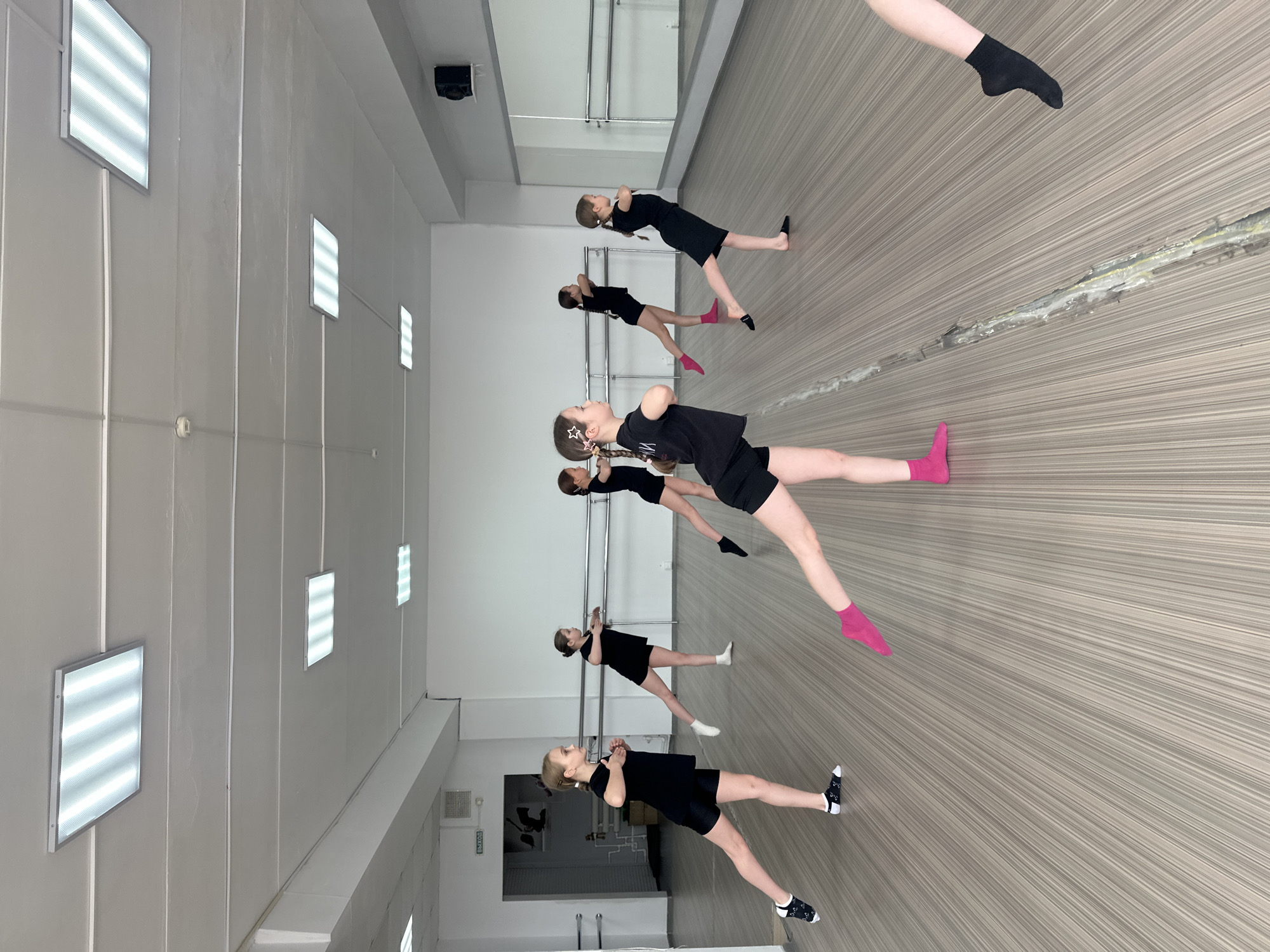 In what appears to be a dance studio located in Votkinsk, Russia, six girls in black dancewear are practicing a dance routine. They are standing in a line, with arms crossed over their chests, and one leg extended to the side. Their sock colors vary, with some wearing black, white, or pink socks. The studio has mirrors along one wall, ballet barres, and fluorescent lights on the ceiling. The floor is a light brown color. The photo appears to have been taken at an angle, and then rotated for viewing.