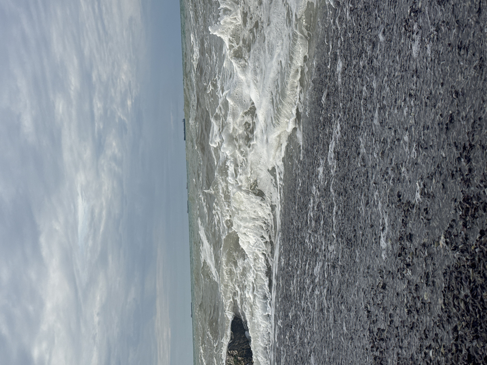 The image depicts a choppy sea with gray, overcast skies. Waves are breaking on a dark, pebbly shore, with some foam visible in the water. In the distance, there are several ships on the horizon. A dark rock is visible on the left side of the frame, partially submerged in the water. The overall impression is of a slightly stormy or windy day at the coast. The location is the Caspian Sea coast of Aktau, Kazakhstan.