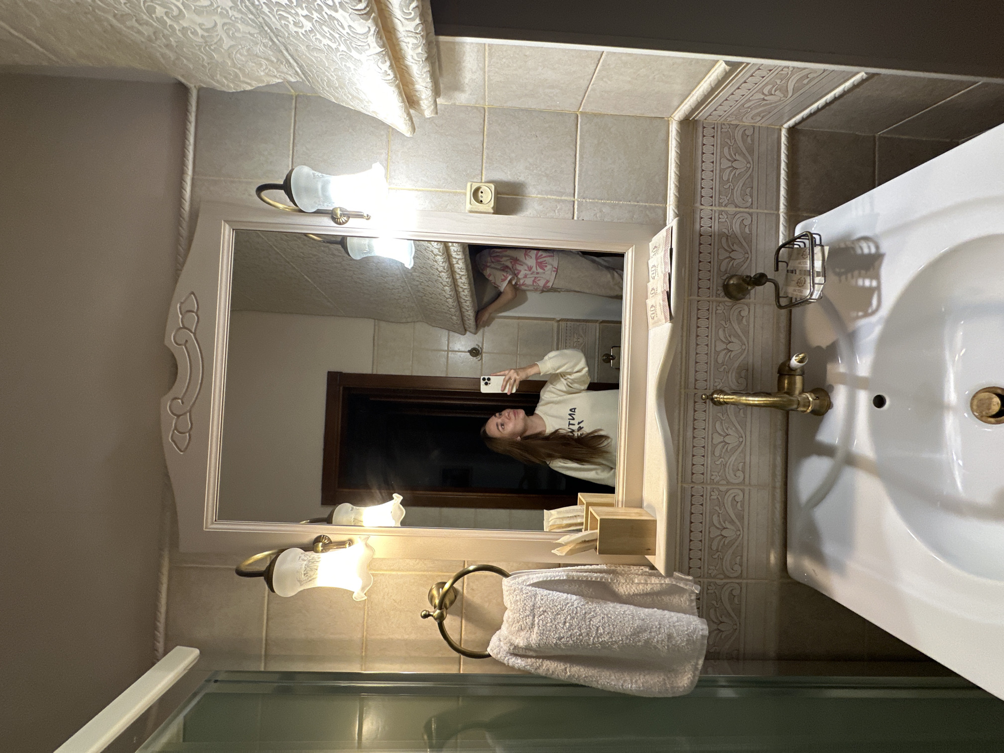 The image depicts a bathroom interior, likely in Khvalynsk, Russia, with a young woman taking a selfie in a mirror. She is wearing a light-colored sweatshirt with text on it. In the reflection of the mirror, another person is visible, seemingly adjusting window blinds. The bathroom features beige tiled walls, a white sink with a brass faucet, a towel rack with a white towel, and decorative wall sconces. Small packets, possibly toiletries, are arranged on a shelf above the sink. The overall impression is that of a standard hotel or guesthouse bathroom.