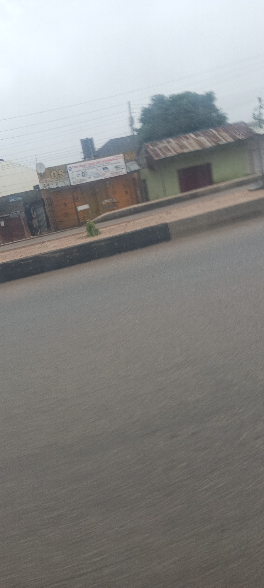 The image presents a roadside scene in Jos, Nigeria, captured from a moving perspective, evidenced by the motion blur across the road surface in the foreground. The weather appears overcast, with a diffused, pale sky.
In the immediate foreground, a grey asphalt road spans the lower portion of the frame. It meets a dark-colored curb topped with a lighter strip, separating it from the establishments along the road. A small cluster of green plants grows on this curb.
Along the roadside, a series of structures come into view. On the left, a building is partially visible, featuring a satellite dish. Next to it is a commercial premises with a large, closed, brownish-orange metal gate or wall. Above this gate, a prominent white rectangular sign displays "DON-CHUKS EXCELLENT AUTOMOBILE LTD" in clear lettering. Below this, smaller text is partially discernible, including "The Quality..." and "Tyre Sales", alongside some graphics. To the left of the main business name, the letters "OS" are visible. A black water tank is situated on the roof behind the sign.
Further to the right stands a light green-painted building with a dark, possibly red, door. Its roof is constructed from corrugated metal sheets, showing signs of rust. Behind the buildings, a large tree with dense green foliage and utility poles with overhead wires are visible against the pale sky. A faint, blurry dark figure, possibly a person, can be seen near the left side of the large brown gate. The scene depicts a typical urban or semi-urban street, with no specific event unfolding beyond the general activity of the street.