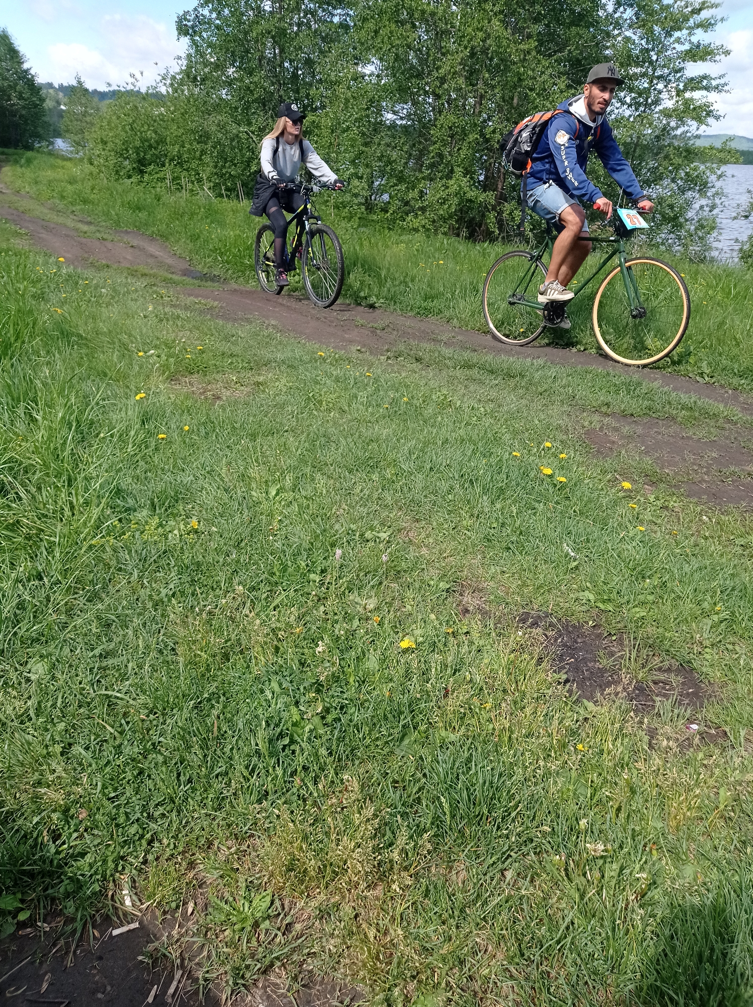 Two individuals are riding bicycles on a dirt road adjacent to a grassy area, with a body of water and trees visible in the background. The individual on the right is a man wearing a blue jacket, denim shorts, a baseball cap, and carrying a backpack. He is riding a green bicycle with tan-colored tires and has a number visible on the front. The individual on the left is a woman wearing a baseball cap, a light-colored long-sleeved shirt, dark leggings, and riding a dark-colored mountain bike. The location appears to be a rural or park area within Degtyarsk, Russia.