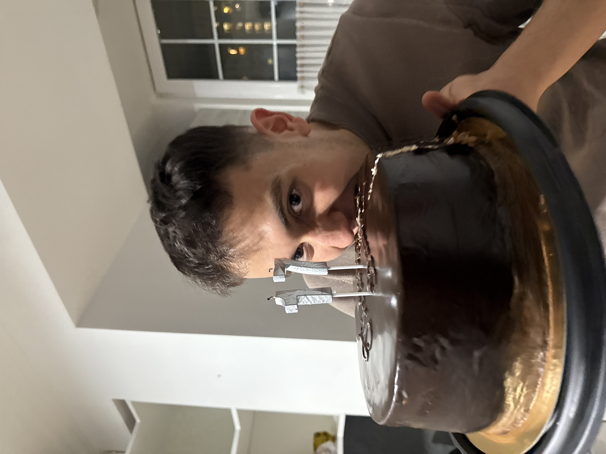A man with short, dark hair and light skin holds a chocolate frosted cake in his right hand. The cake has two number-shaped candles on top that appear to be covered in aluminum foil. The numbers are not clear, but might be a '2' and a '7'. The background shows a window with a view of a city skyline at night. Considering the architectural style of the buildings and the location of the person, it is likely that the person is in the city of Yerevan, Armenia. The room appears to be indoors, possibly an apartment, with white walls and a partial view of a shelf or cabinet. The man's face is angled towards the cake. He is wearing a brown t-shirt.